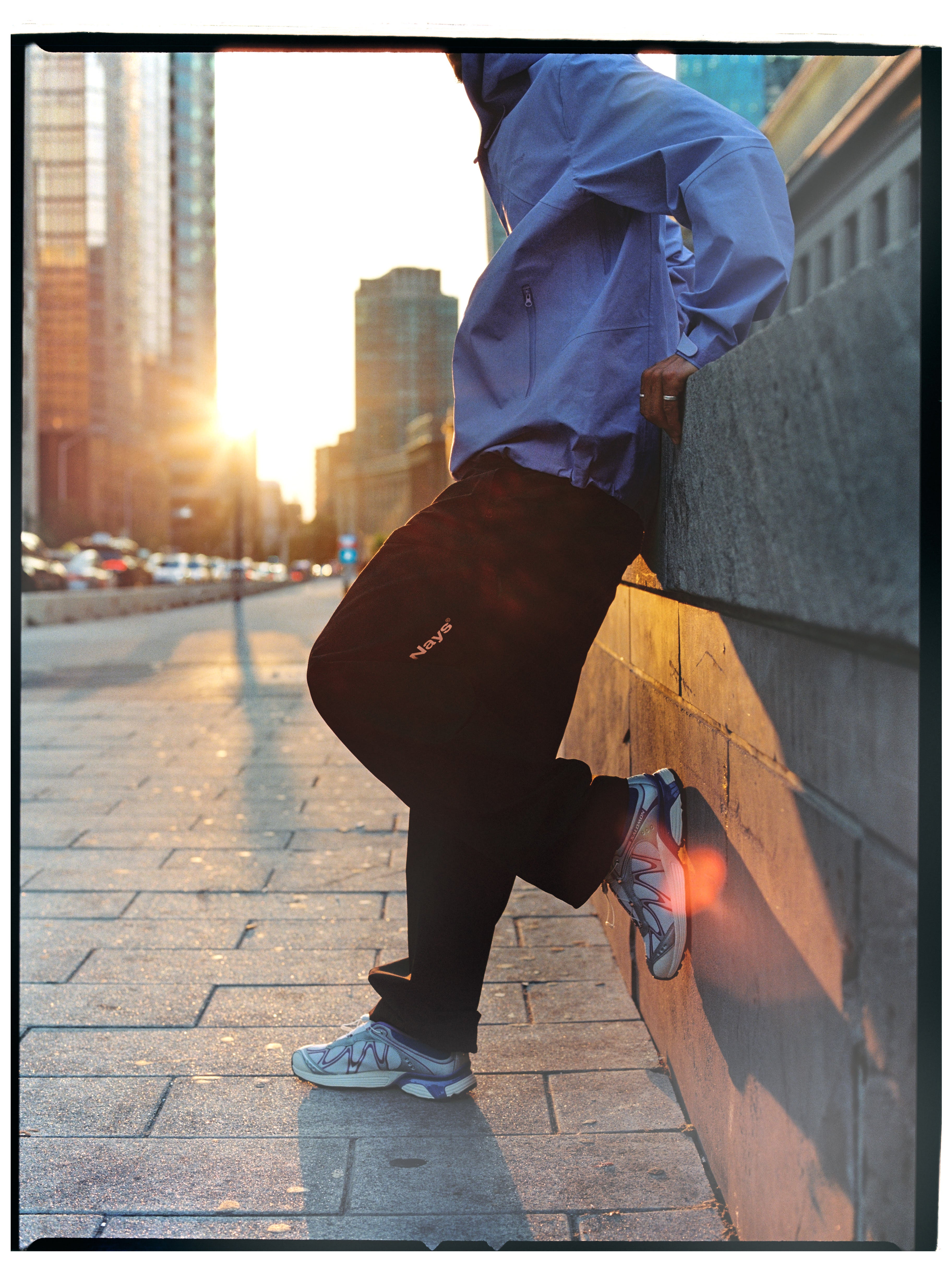Seitenansicht der lilafarbenen NAYS Rain Jacket 2.0 mit schwarzer Nays Hose im Sonnenuntergang in Toronto.
