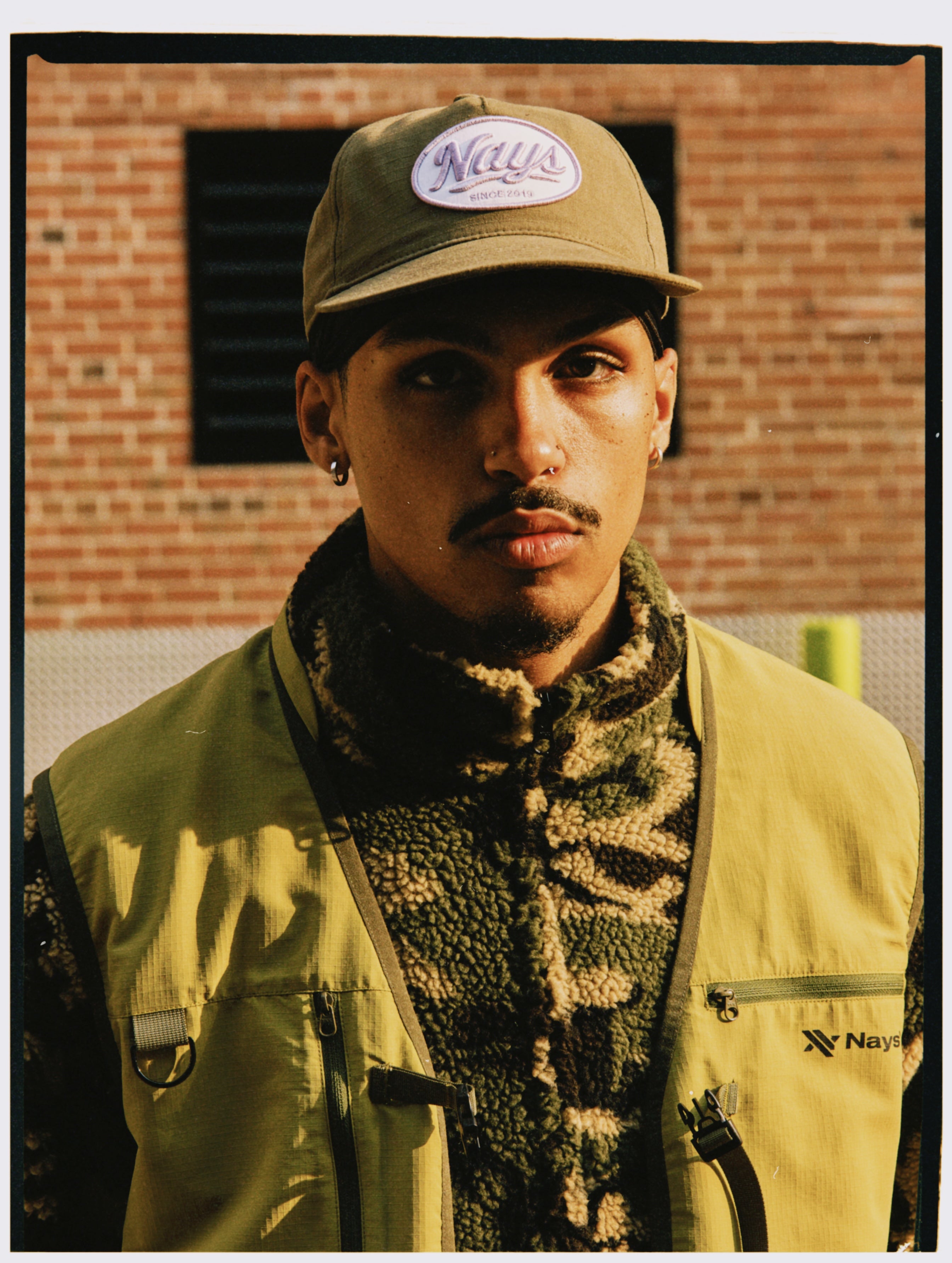 Model mit olivgrüner NAYS Cap, Camouflage Fleece und Weste vor einer Backsteinwand in Toronto.