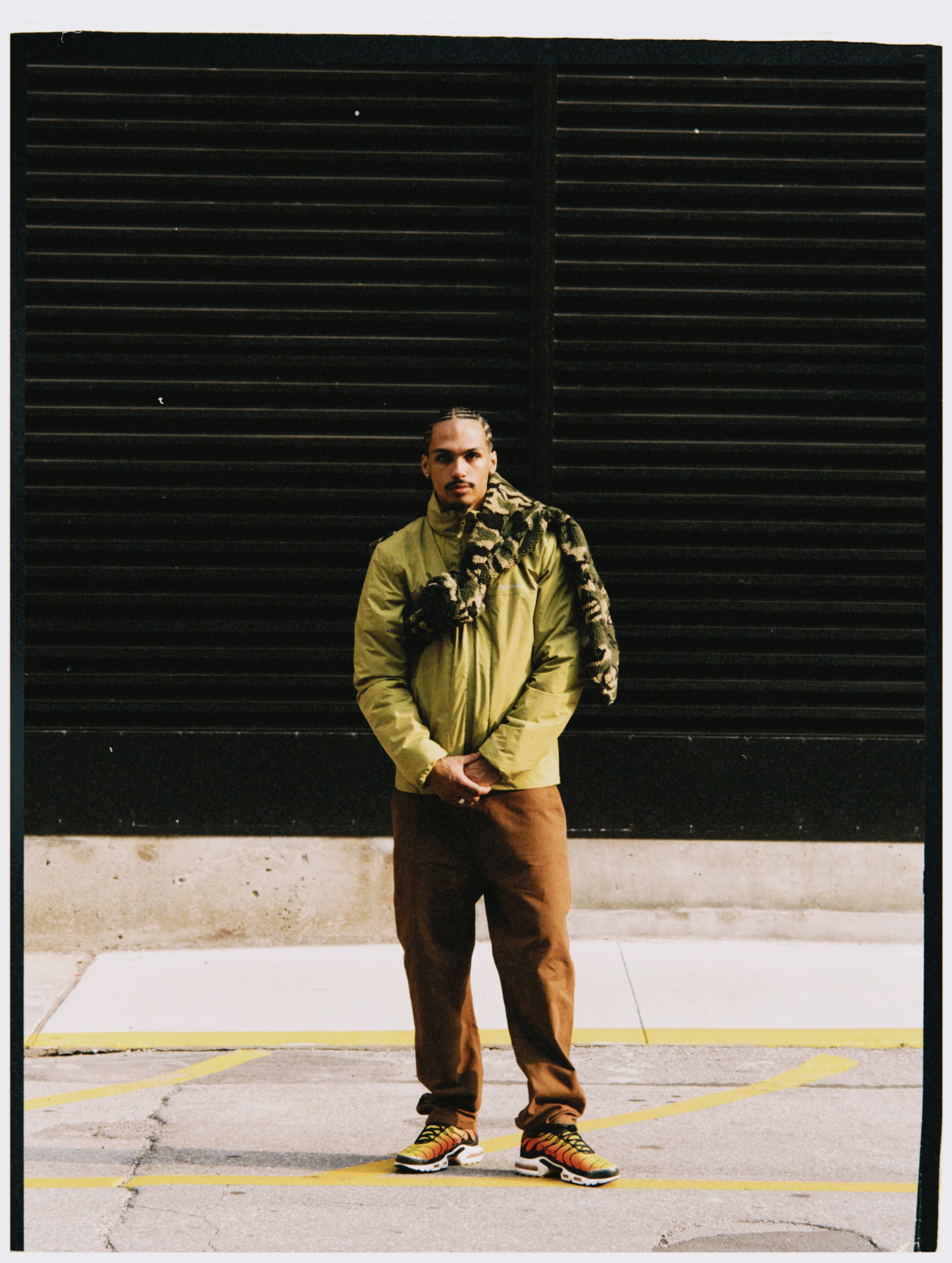 Model in olivgrüner NAYS Jacke, brauner Hose und Camouflage-Fleece vor einer schwarzen Industriefassade.