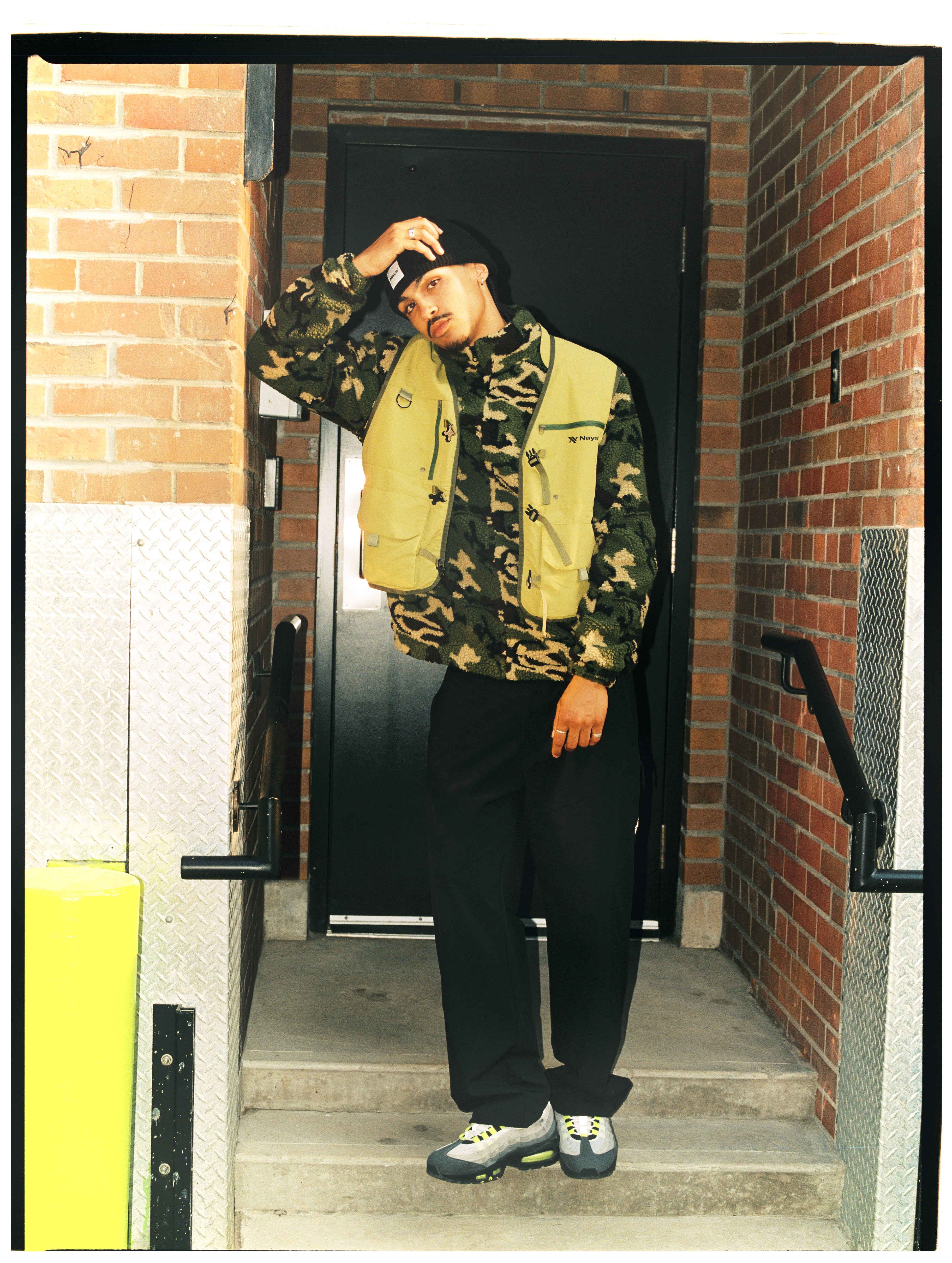 Model in NAYS Camouflage-Fleece und olivgrüner Utility-Weste vor einem Backsteingebäude in Toronto.