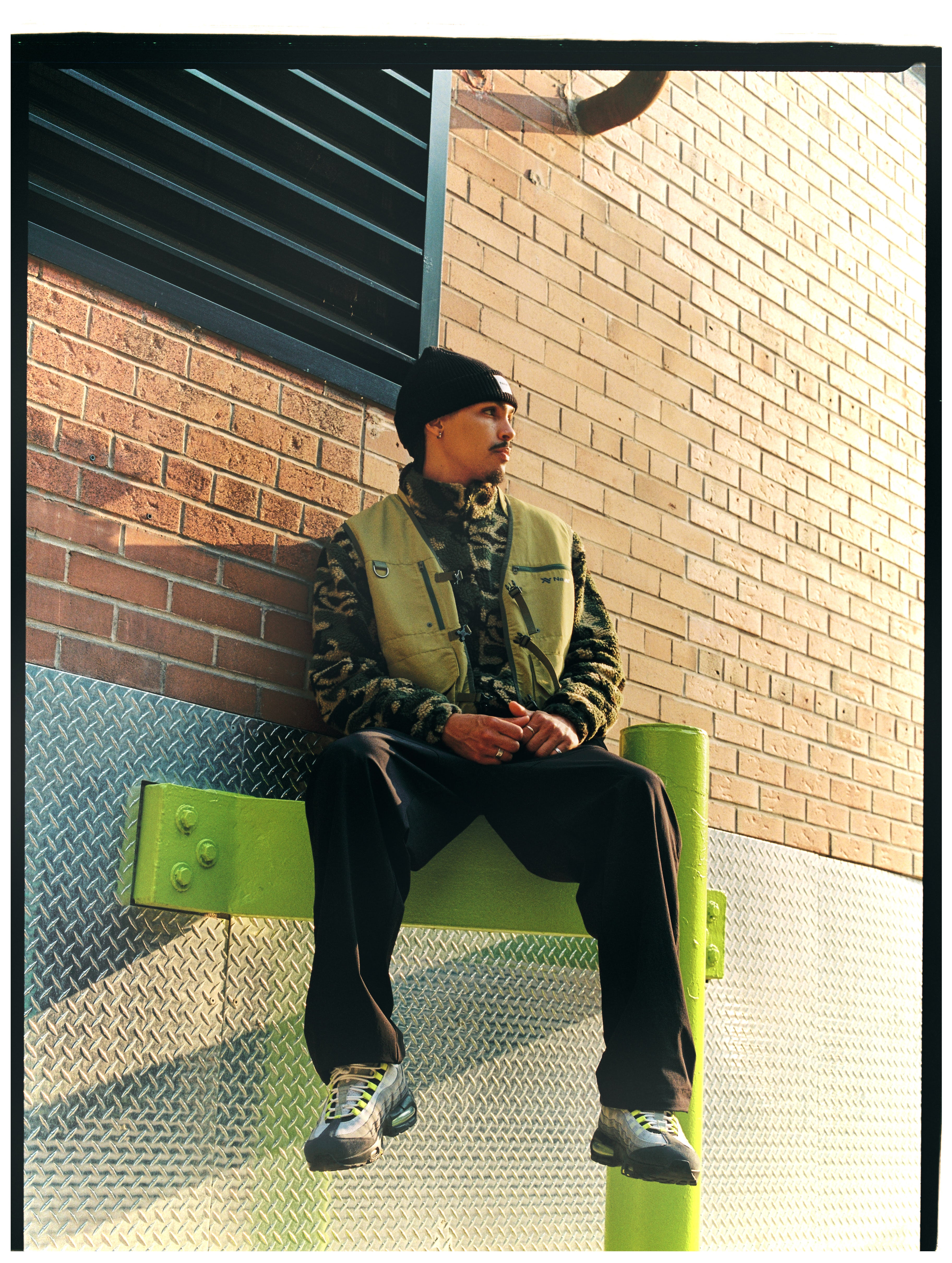 Model mit NAYS Utility-Weste und Camouflage-Fleece, sitzend vor einer Backsteinfassade in Toronto.