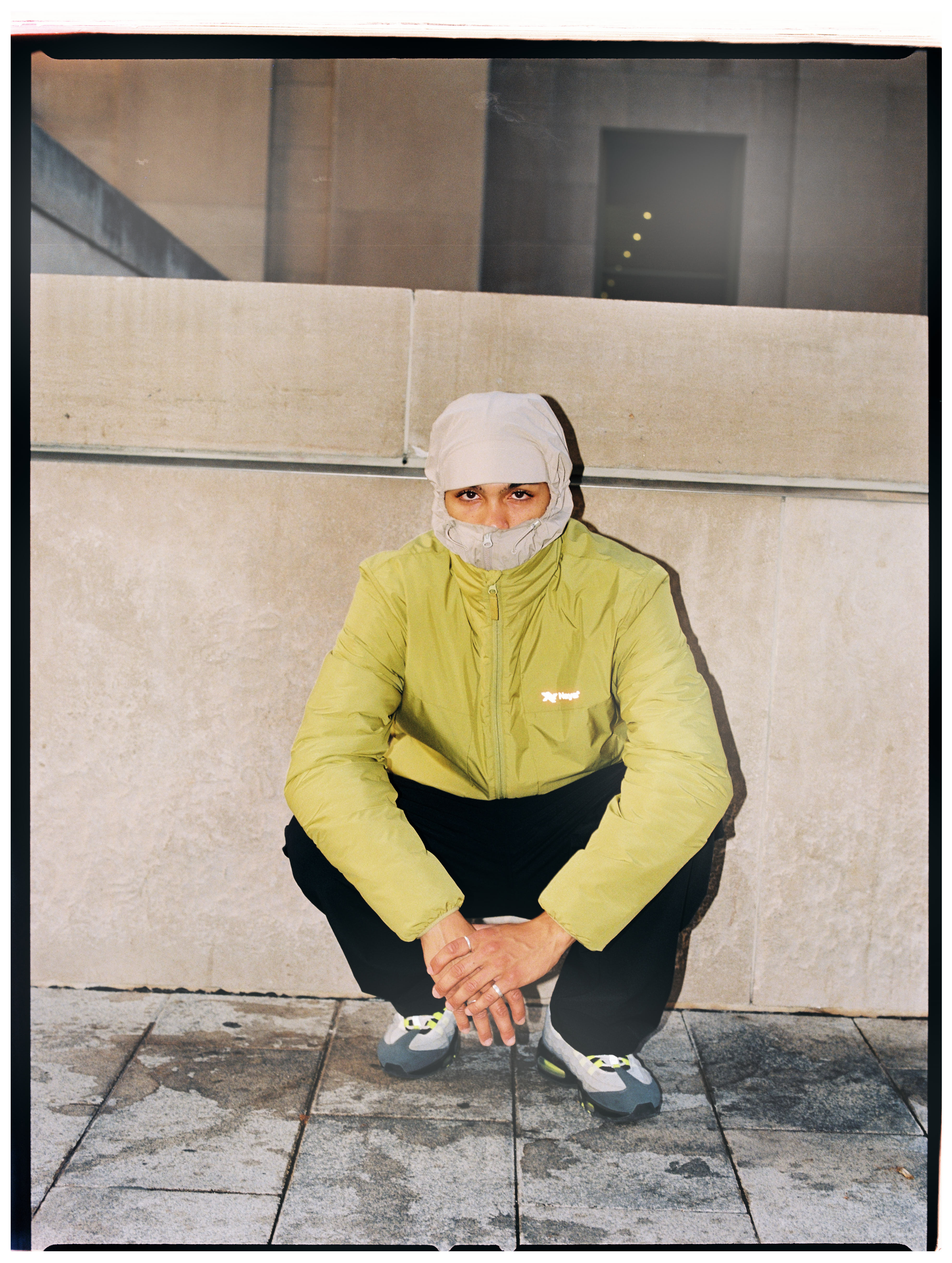 Model in hellgrüner NAYS Jacke mit hochgezogenem Gesichtsschutz, schwarzer Hose und Sneakers vor einer Betonwand.