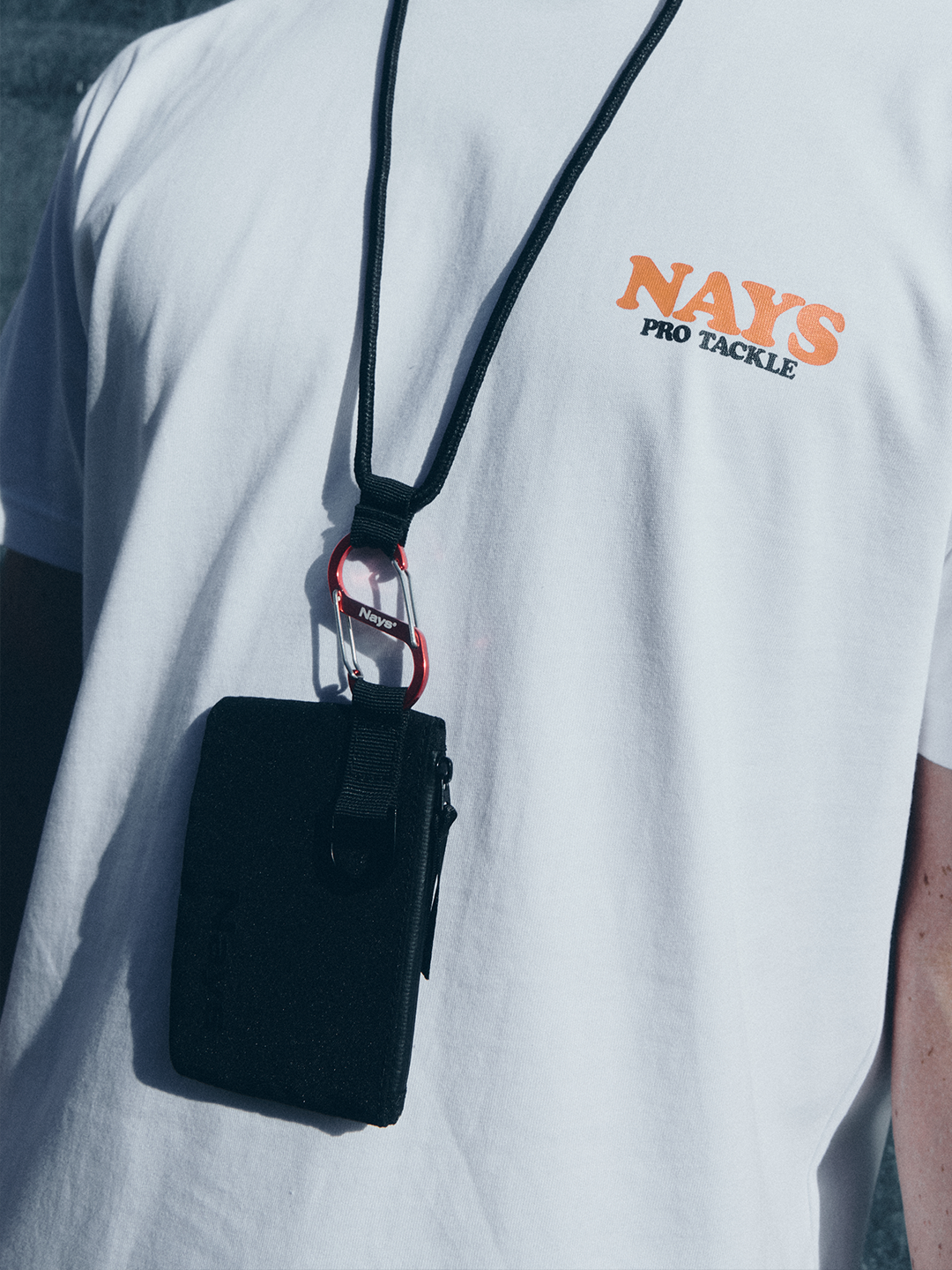 Model in hellem NAYS College Print T-Shirt mit minimalistischer Grafik und NAYS Neck Pouch vor cleanem urbanem Hintergrund. 