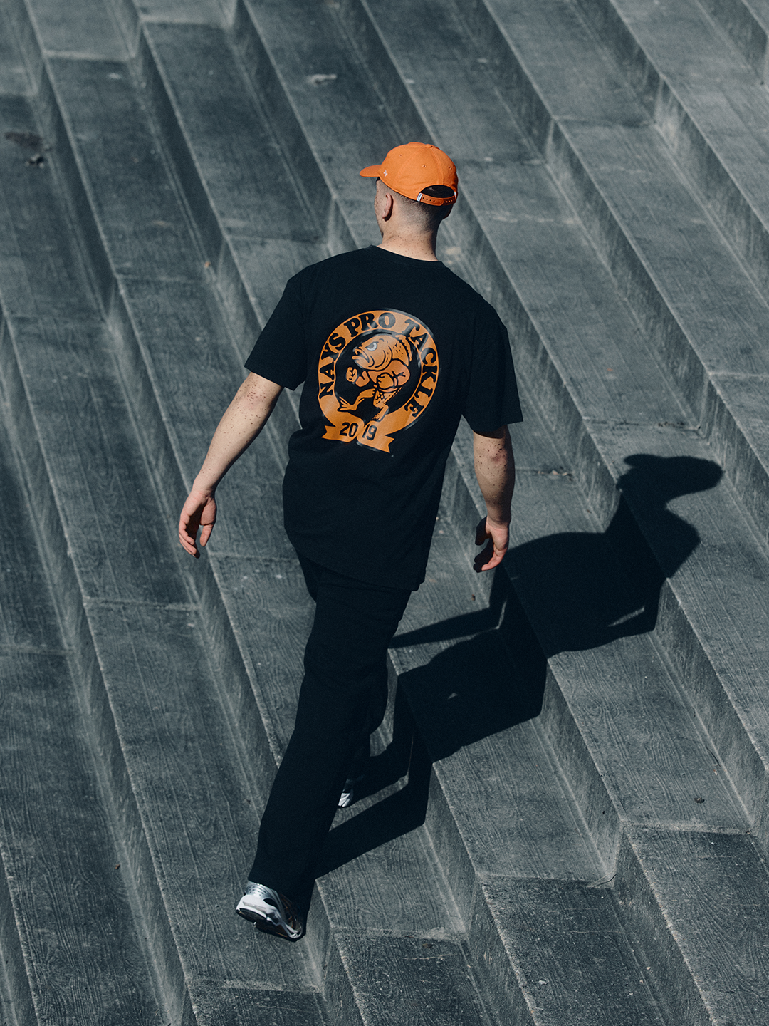 Model in schwarzem NAYS College Print T-Shirt und orangener Fishing Club Cap im urbanen Setting des Velodrom Berlin.