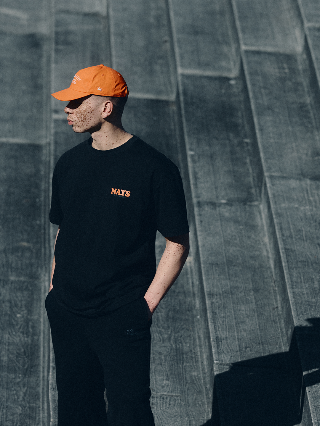Model in schwarzem NAYS College Print T-Shirt und orangener Fishing Club Cap im urbanen Setting des Velodrom Berlin.