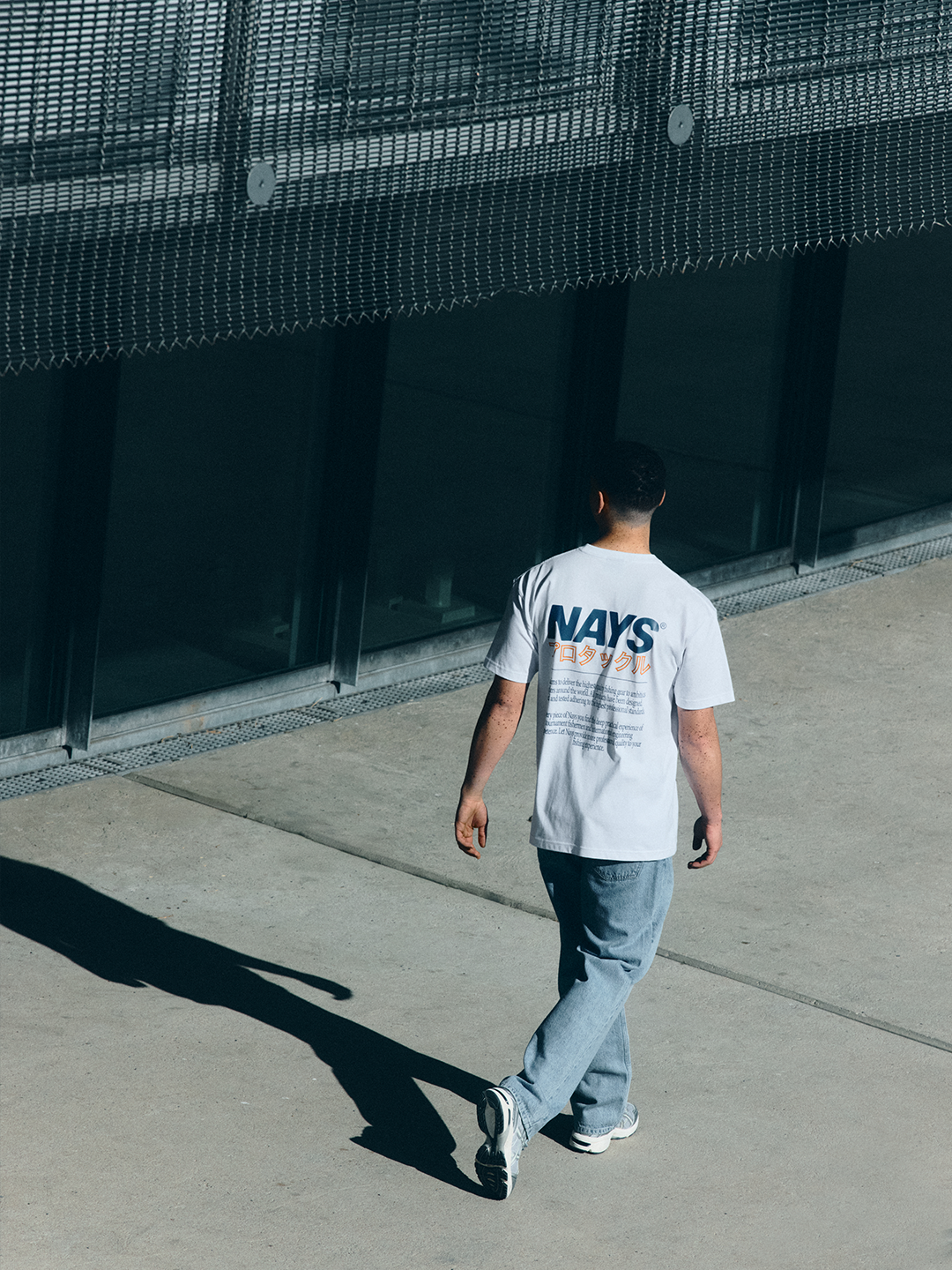 Model in hellem NAYS Pro Tackle International T-Shirt vor moderner Architektur mit cleanem, sportlichem Look.