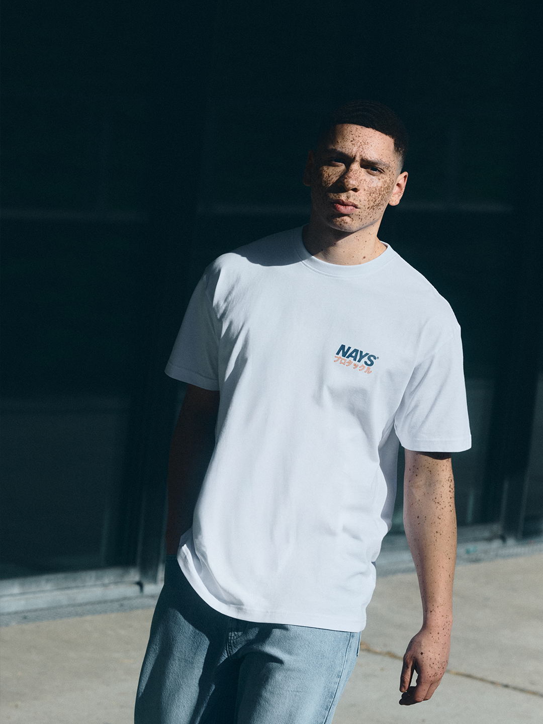 Model in hellem NAYS Pro Tackle International T-Shirt vor moderner Architektur mit cleanem, sportlichem Look.