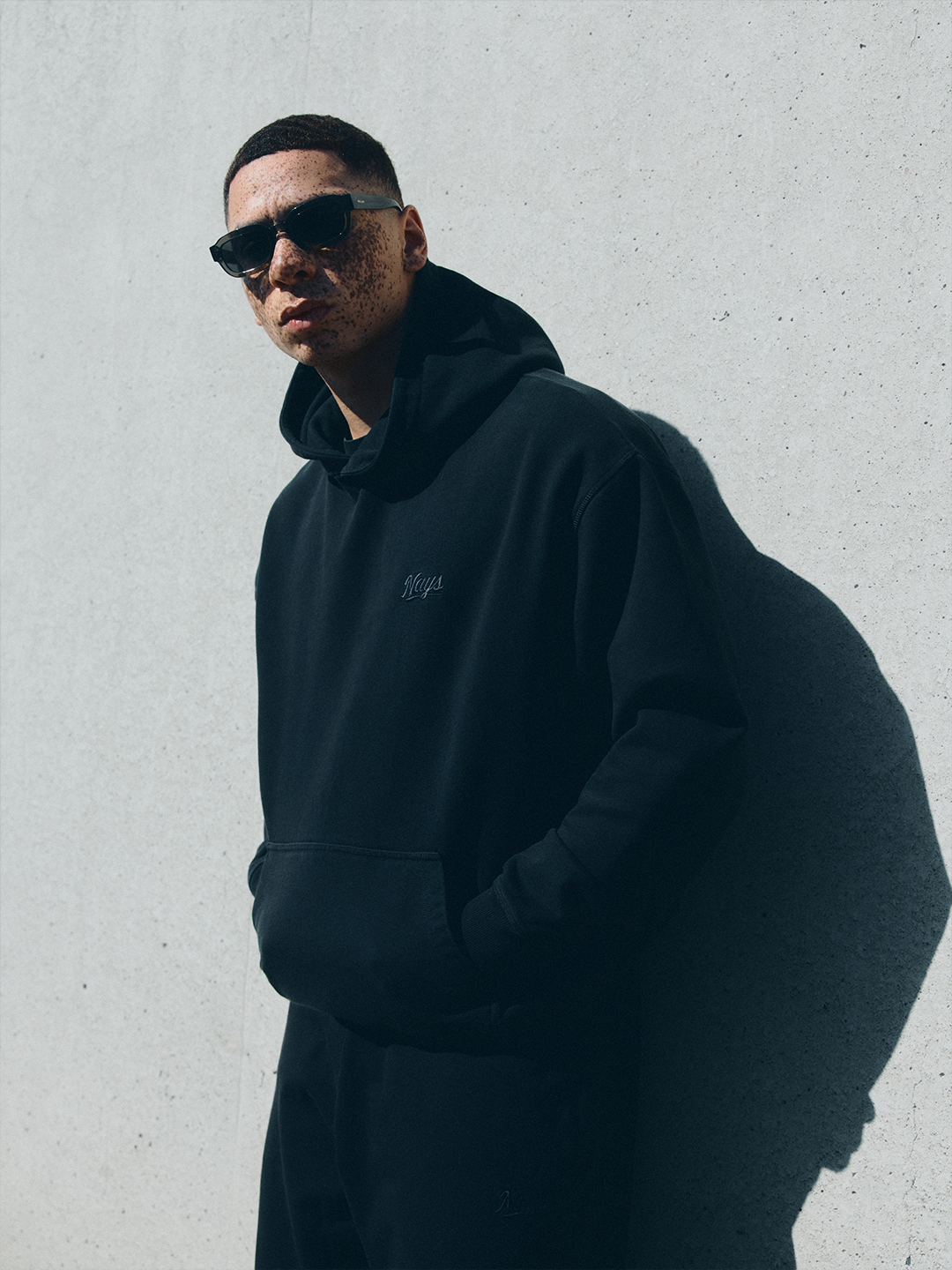 Model in schwarzem NAYS New Script Hoodie und Sonnenbrille vor minimalistischer Betonwand im urbanen Setting des Velodrom Berlin.