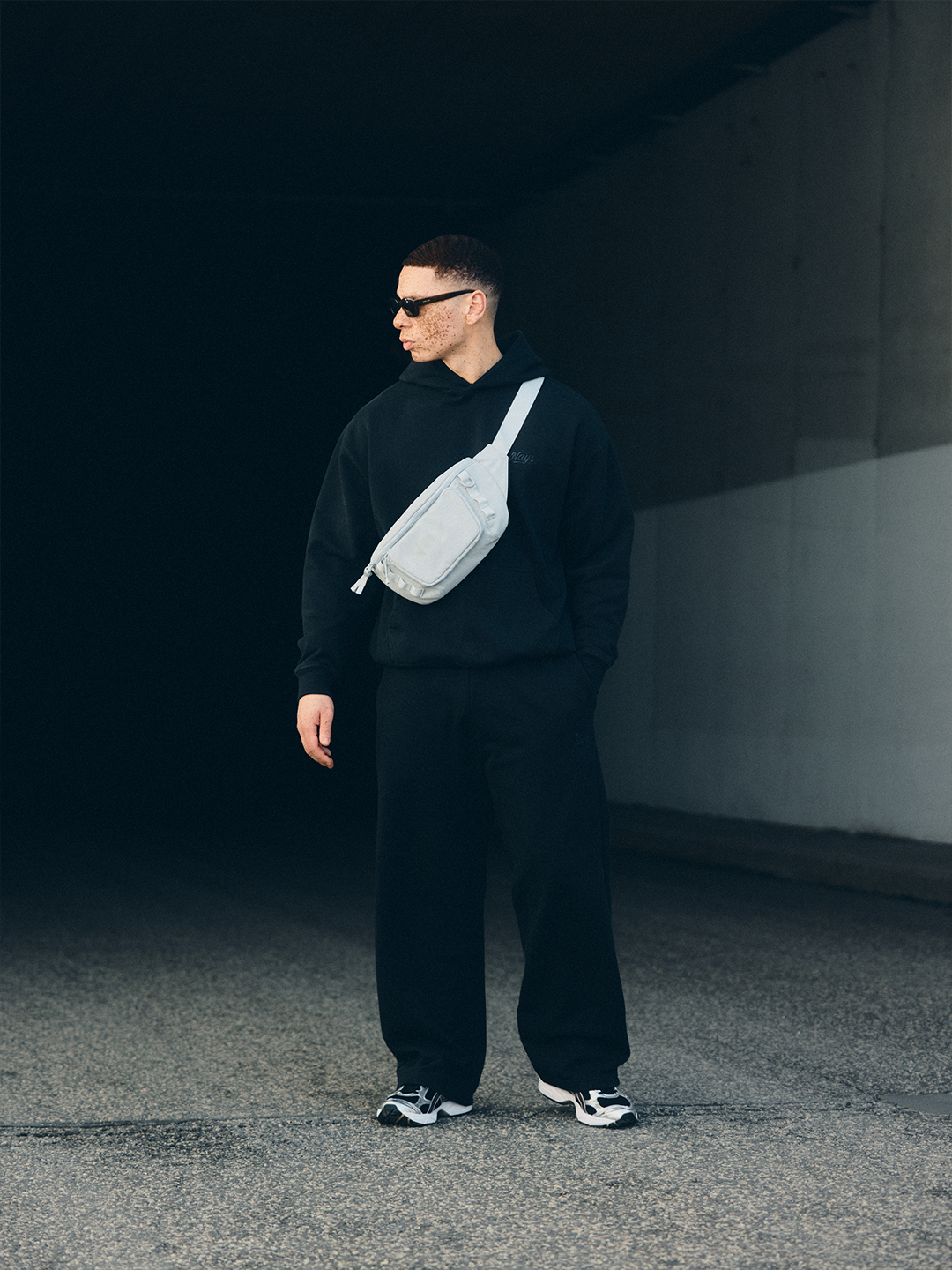 Model in schwarzem NAYS New Script Hoodie, Straight Leg Pants mit heller Nays Lightweight Cross Body Bag und Sonnenbrille vor urbaner Betonarchitektur im Velodrom Berlin.