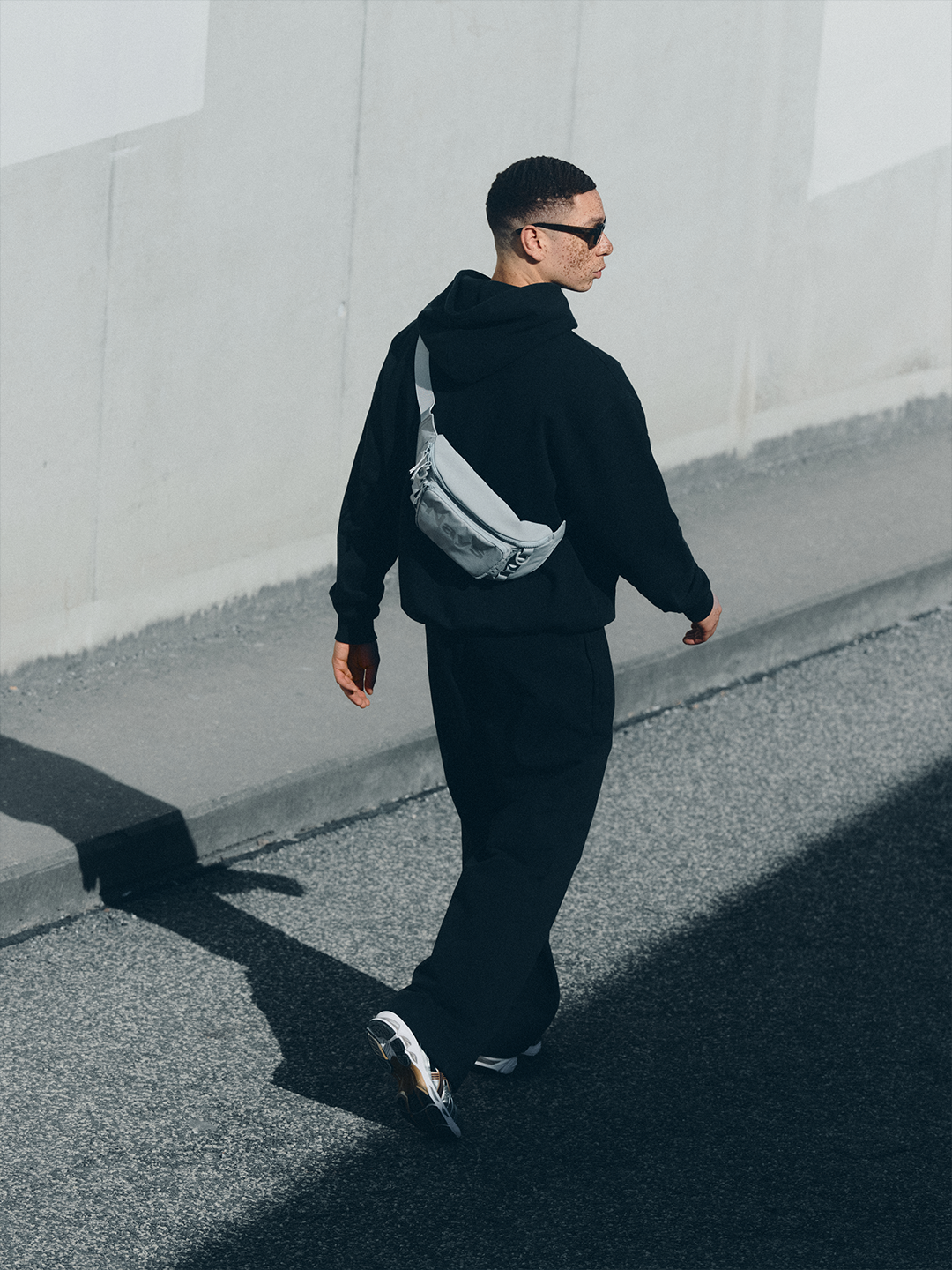 Model in schwarzem NAYS New Script Hoodie, Straight Leg Pants mit heller Nays Lightweight Cross Body Bag und Sonnenbrille vor urbaner Betonarchitektur im Velodrom Berlin.