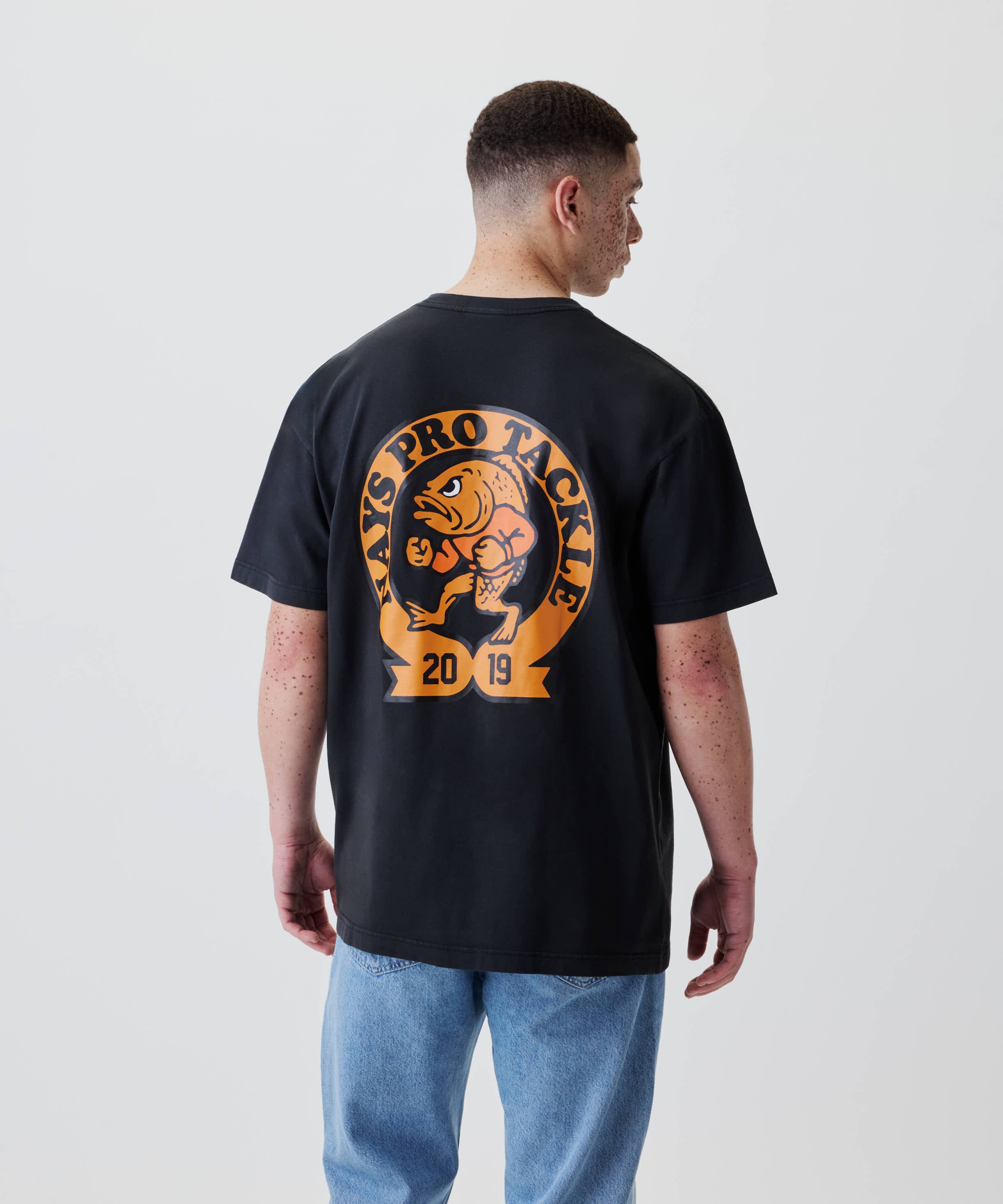 Rückansicht des Nays College Print Tee mit großem orangenem Fisch-Print und AXS PRO TACKLE 2019 Schriftzug auf der Rückseite, getragen von einem jungen Mann.