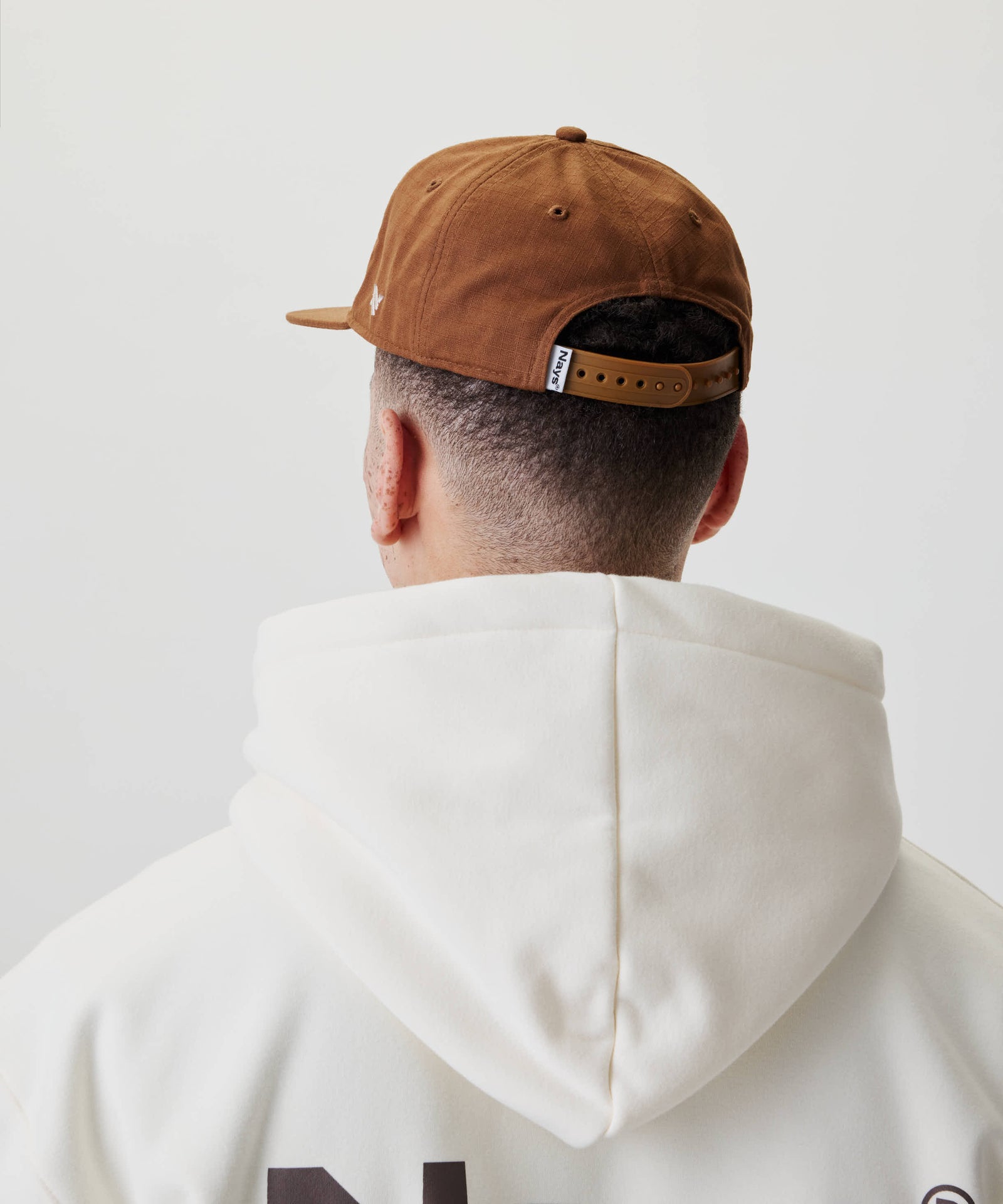 Person von hinten mit einem nach hinten gedrehten Ripstop Script Cap mit verstellbarem Riemen, kombiniert mit einem cremefarbenen Hoodie ohne Kapuze.