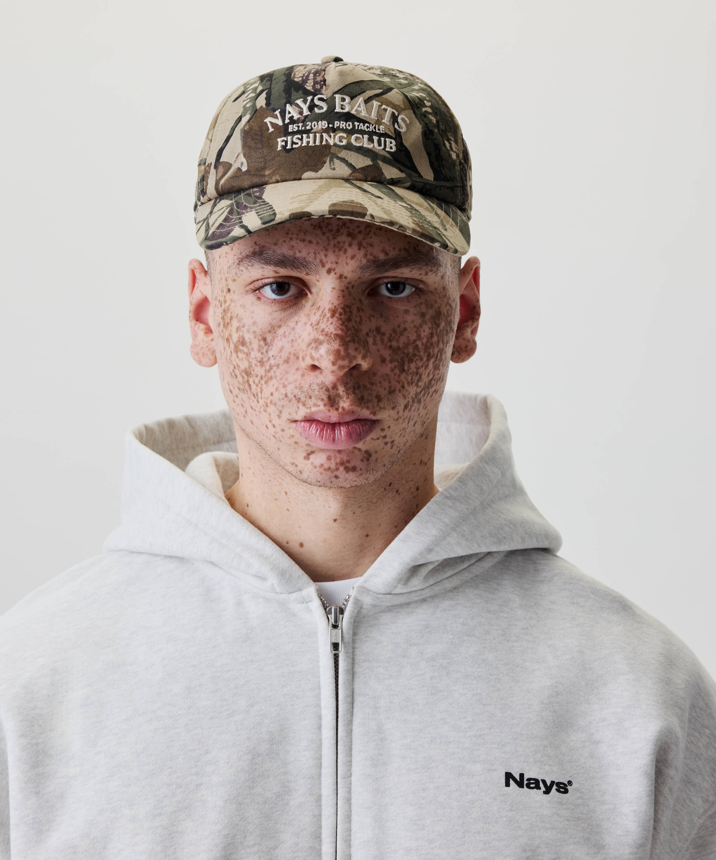 Junge Person mit Nays Fishing Club Cap im Camo-Print, gebogenem Schirm, off-white Stickerei, und grauem Hoodie mit kleinem Nays® Logo, schaut direkt in die Kamera.