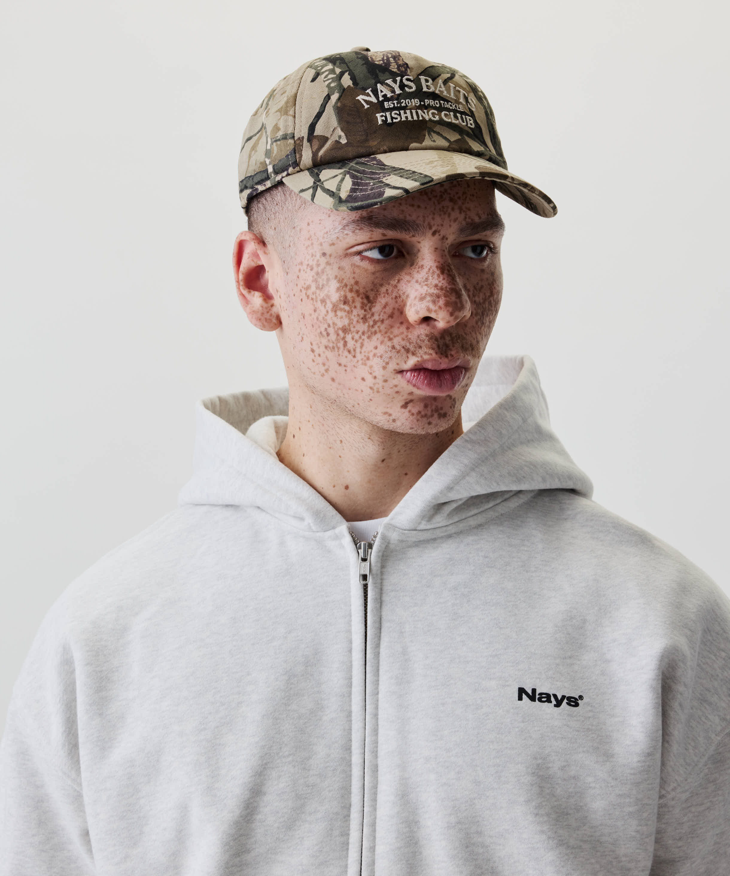 Junger Mann mit Nays Fishing Club Cap im Camo-Print, gebogenem Schirm und Off-white-Stickerei. Trägt grauen Hoodie mit kleinem Nays®-Logo auf der Brust.