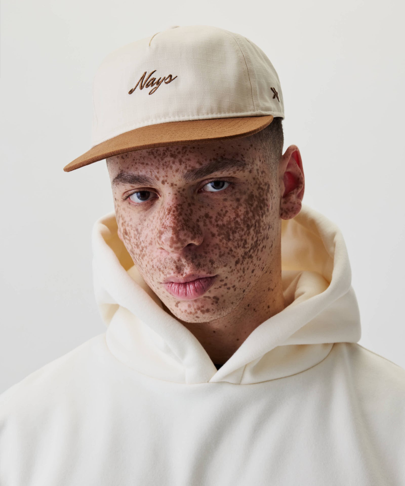 Junge Person mit Sommersprossen trägt die Ripstop Script Cap mit Nays-Stickerei und cremefarbenem Hoodie, vor einem schlichten weißen Hintergrund.