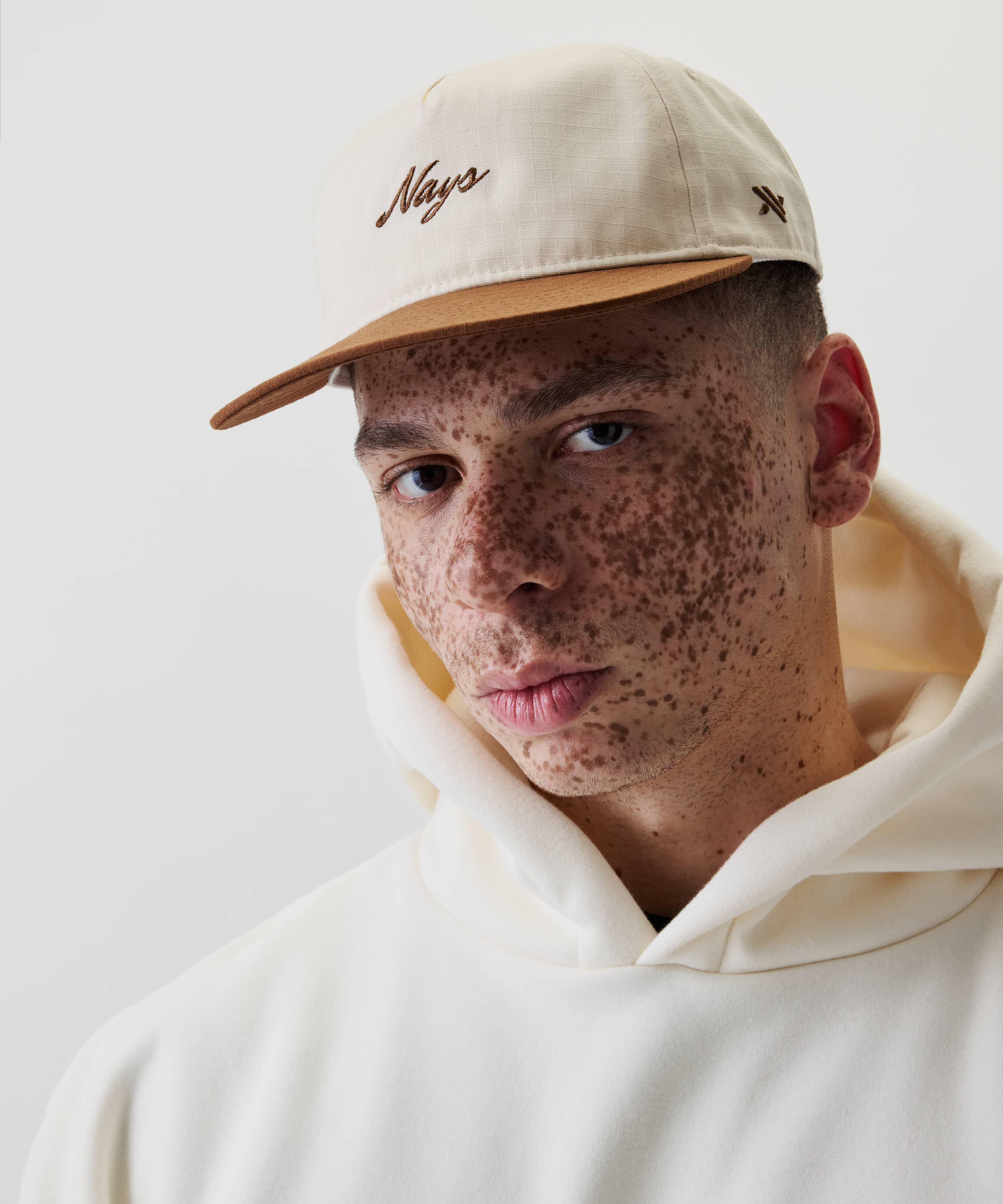 Junge Person mit Sommersprossen trägt Ripstop Script Cap mit braunem Schirm und Nays-Stickerei, kombiniert mit hellen Kapuzenpullover, vor weißem Hintergrund.