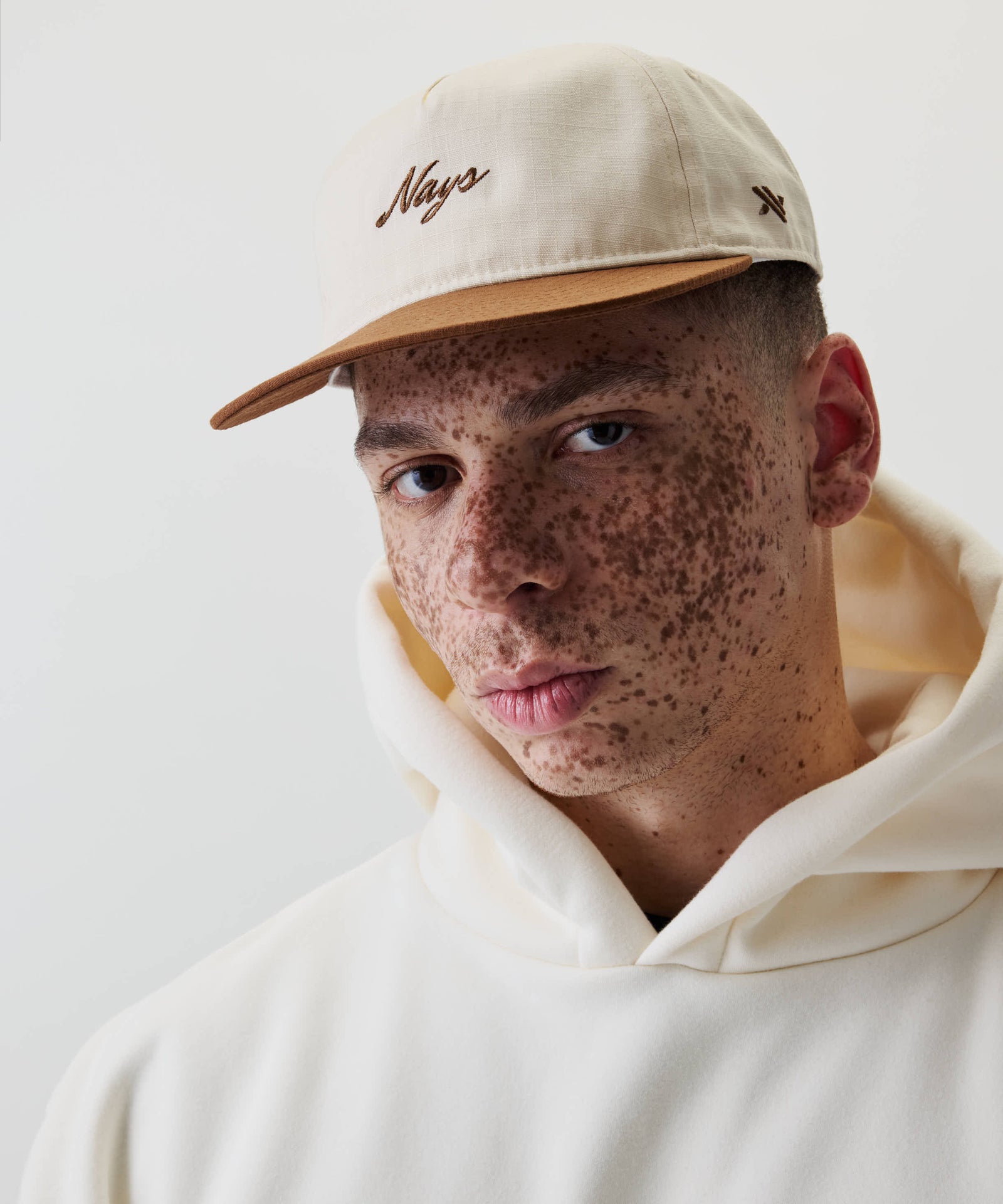 Junge Person mit Sommersprossen trägt Ripstop Script Cap mit braunem Schirm und Nays-Stickerei, kombiniert mit hellen Kapuzenpullover, vor weißem Hintergrund.