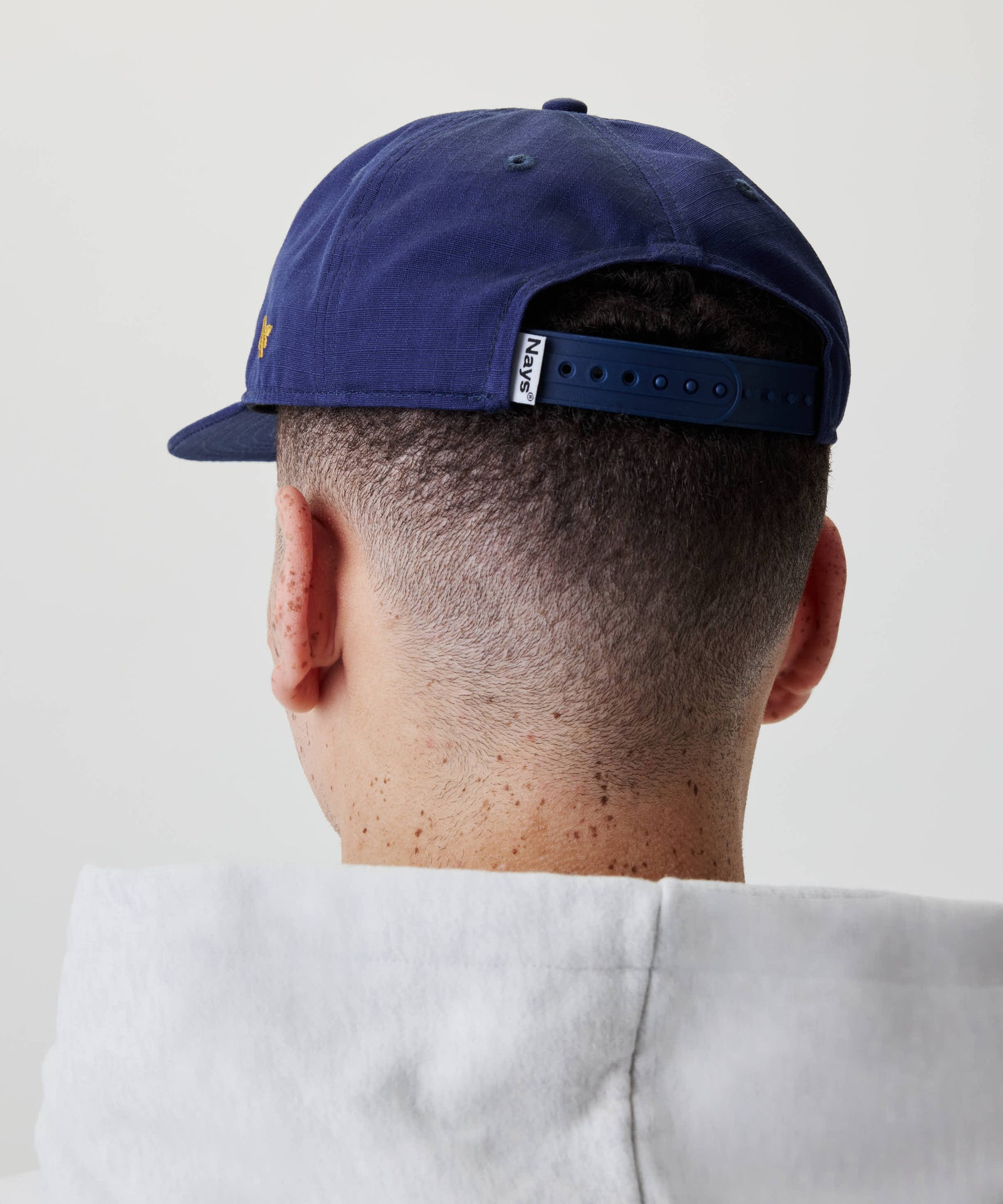Person mit kurzgeschnittenem Haar trägt das Ripstop Script Cap in tiefem Marineblau, mit sichtbarem Nays® Etikett und verstellbarem Riemen, kombiniert mit einem hellen Hoodie.
