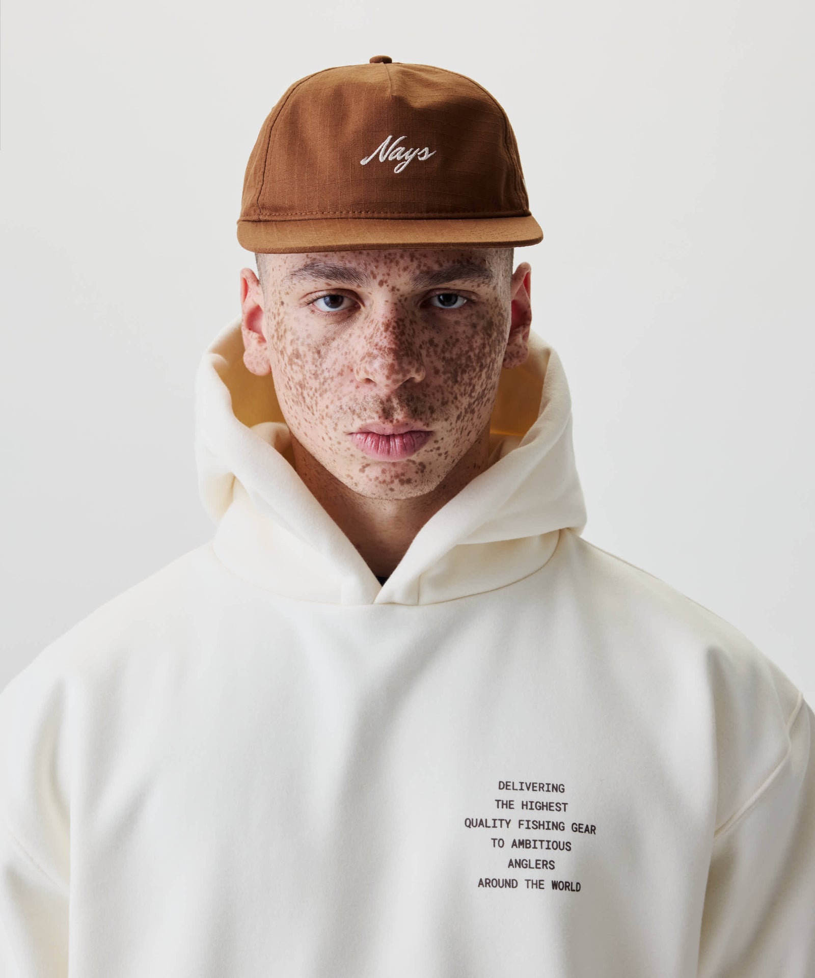 Junge Person trägt ein Ripstop Script Cap mit „Nays“-Stickerei, kombiniert mit einem Hoodie mit Text über hochwertige Angelausrüstung. Studioaufnahme mit weißem Hintergrund.