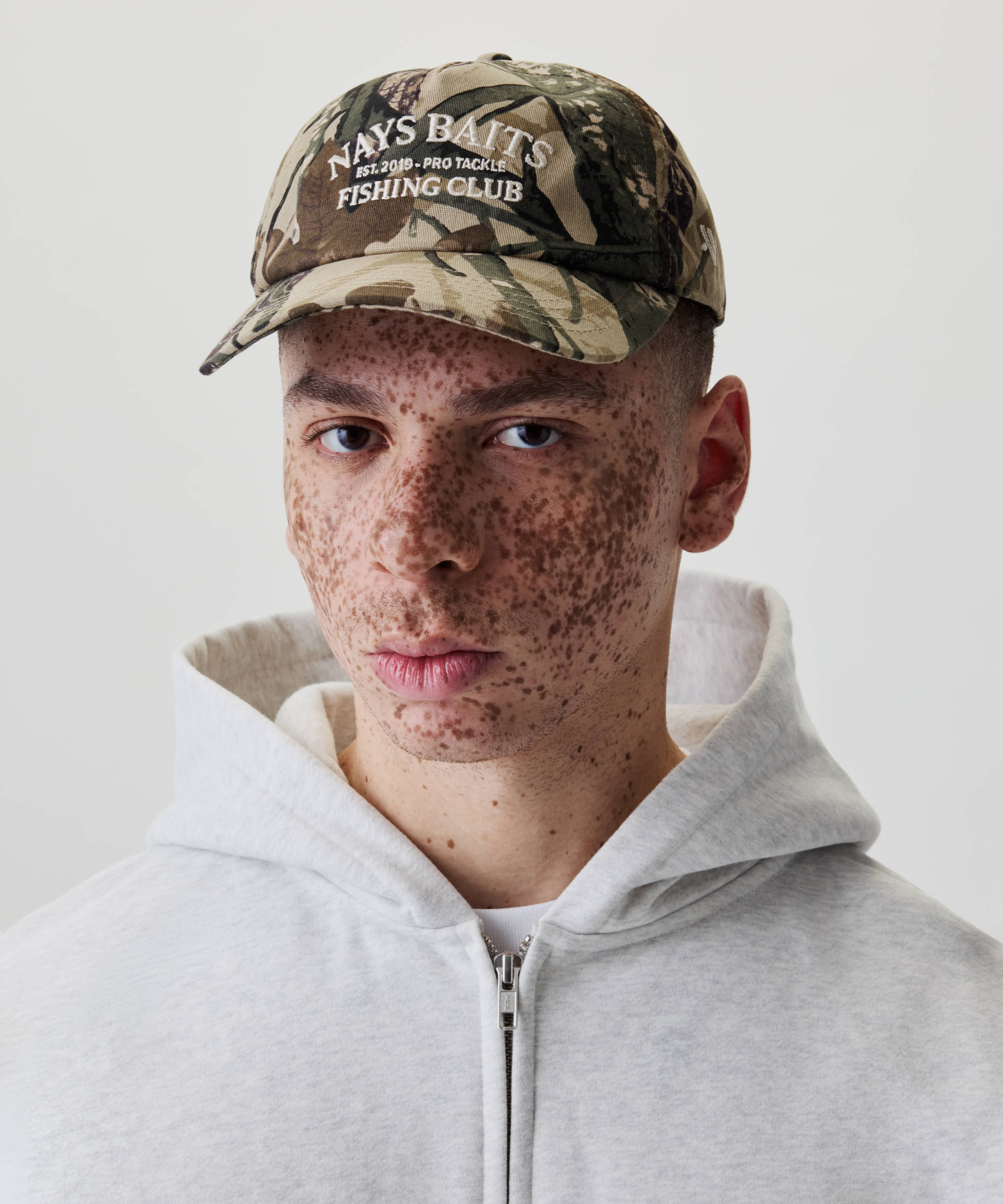 Nahaufnahme einer Person mit Nays Fishing Club Cap im Camo-Print, markanter Off-white-Stickerei und verstellbarem Snapback-Verschluss, kombiniert mit grauem Hoodie.