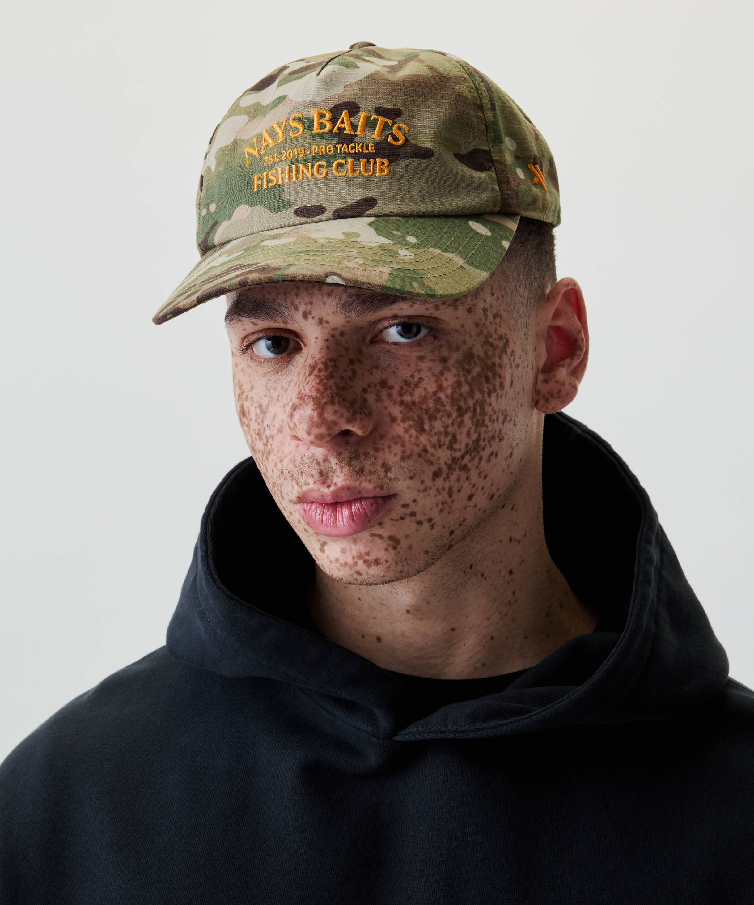 Nahaufnahme einer Person mit Nays Fishing Club Cap im Camo-Print, gelbe Stickerei „PRO TACKLE FISHING CLUB“, verstellbarer Snapback-Verschluss, sportlich-moderner Look.