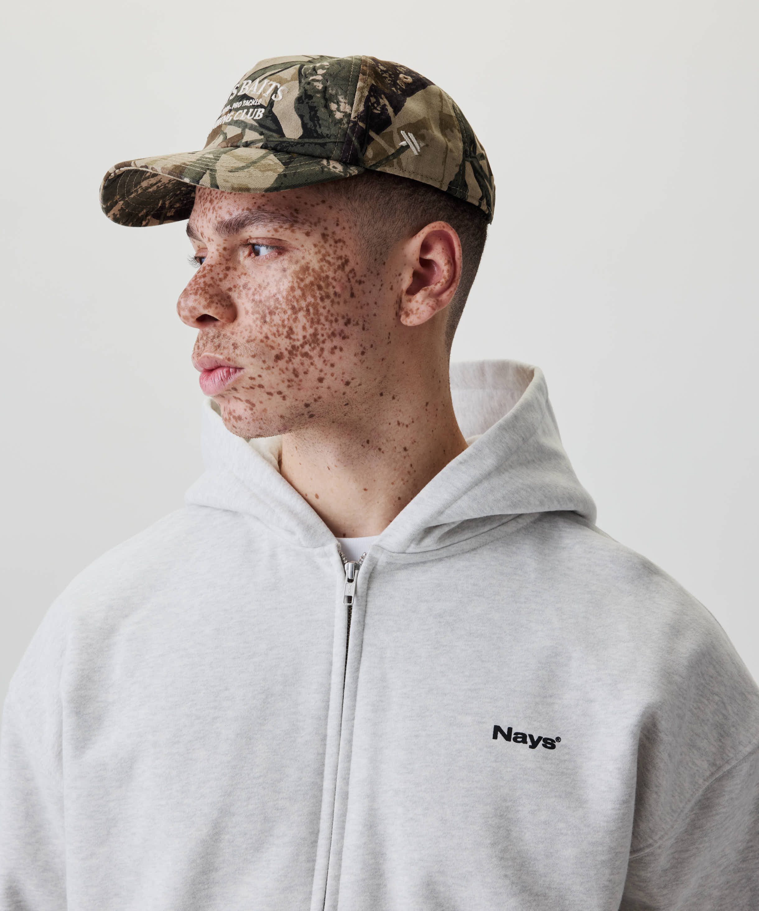 Junger Mann im Profil trägt die Nays Fishing Club Cap mit Camo-Print, gebogenem Schirm und off-white Stickerei, kombiniert mit grauem Hoodie.