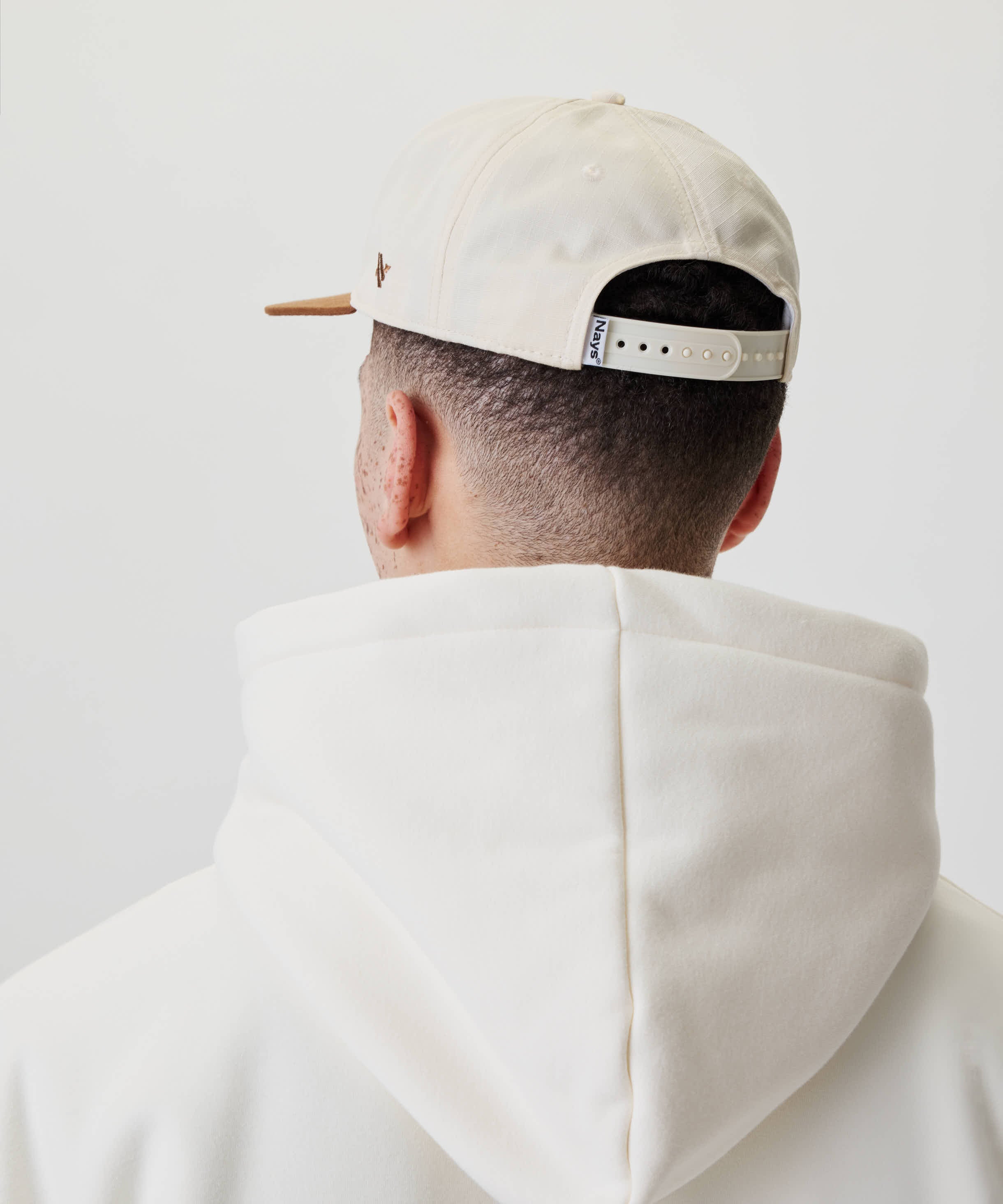 Person von hinten mit sehr kurzem Haar trägt eine Ripstop Script Cap. Die Cap hat einen verstellbaren Riemen mit Nays-Etikett und passt zum Kapuzenpullover.