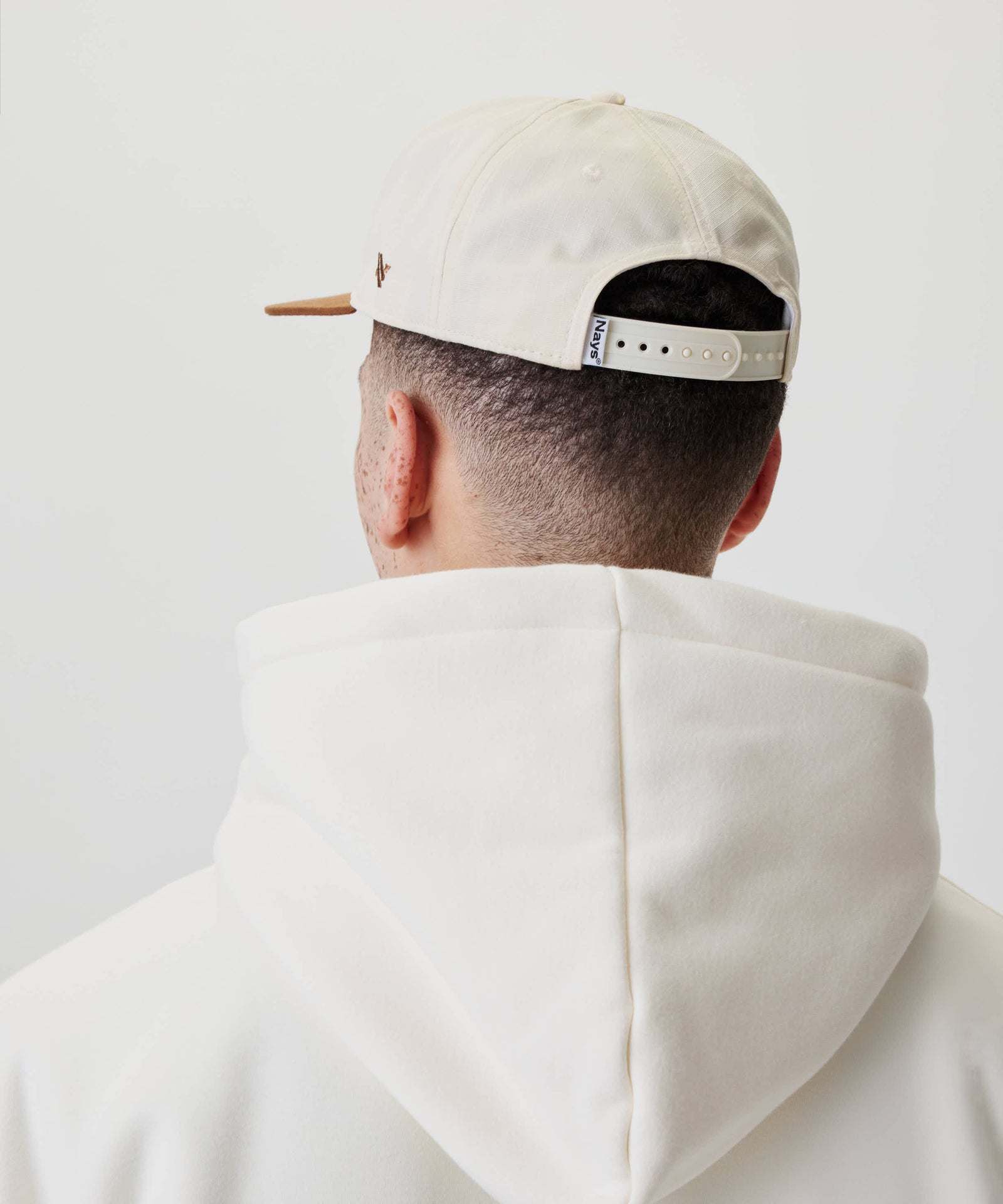 Person von hinten mit sehr kurzem Haar trägt eine Ripstop Script Cap. Die Cap hat einen verstellbaren Riemen mit Nays-Etikett und passt zum Kapuzenpullover.