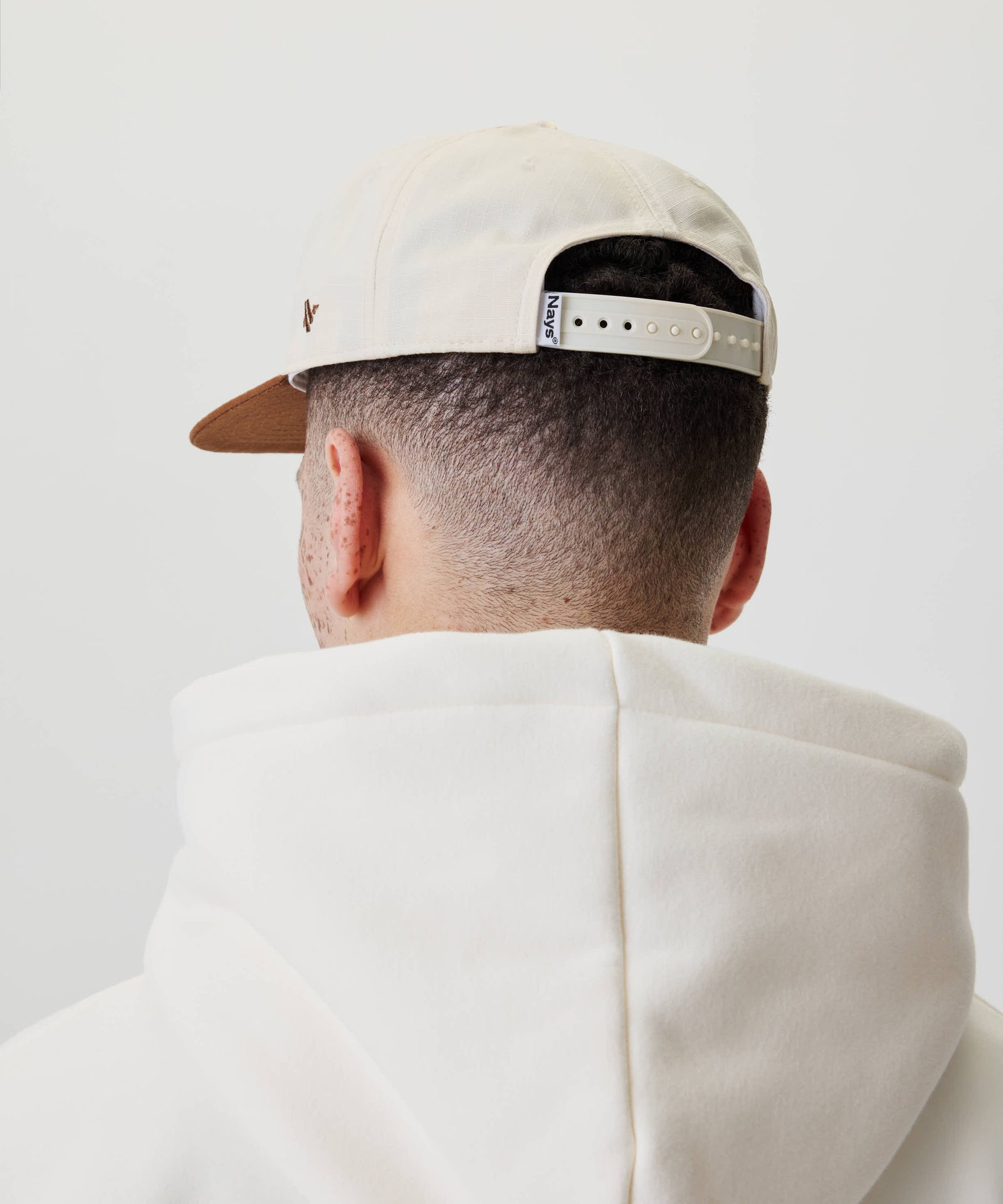 Rückansicht einer Person mit Ripstop Script Cap rückwärts getragen, zeigt verstellbaren Riemen und dezente Sommersprossen auf Nacken und Ohr, trägt cremefarbenen Hoodie.