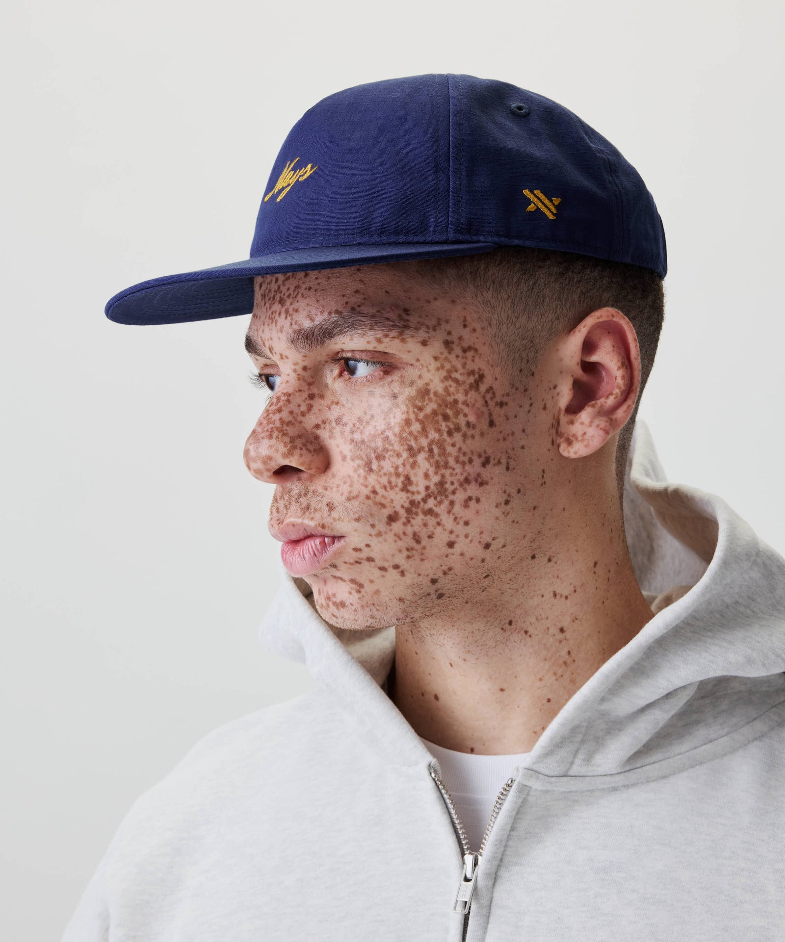 Junger Mann mit Ripstop Script Cap mit flachem Schirm und gelber Stickerei, kombiniert mit einem grauen Hoodie, blickt nach links.