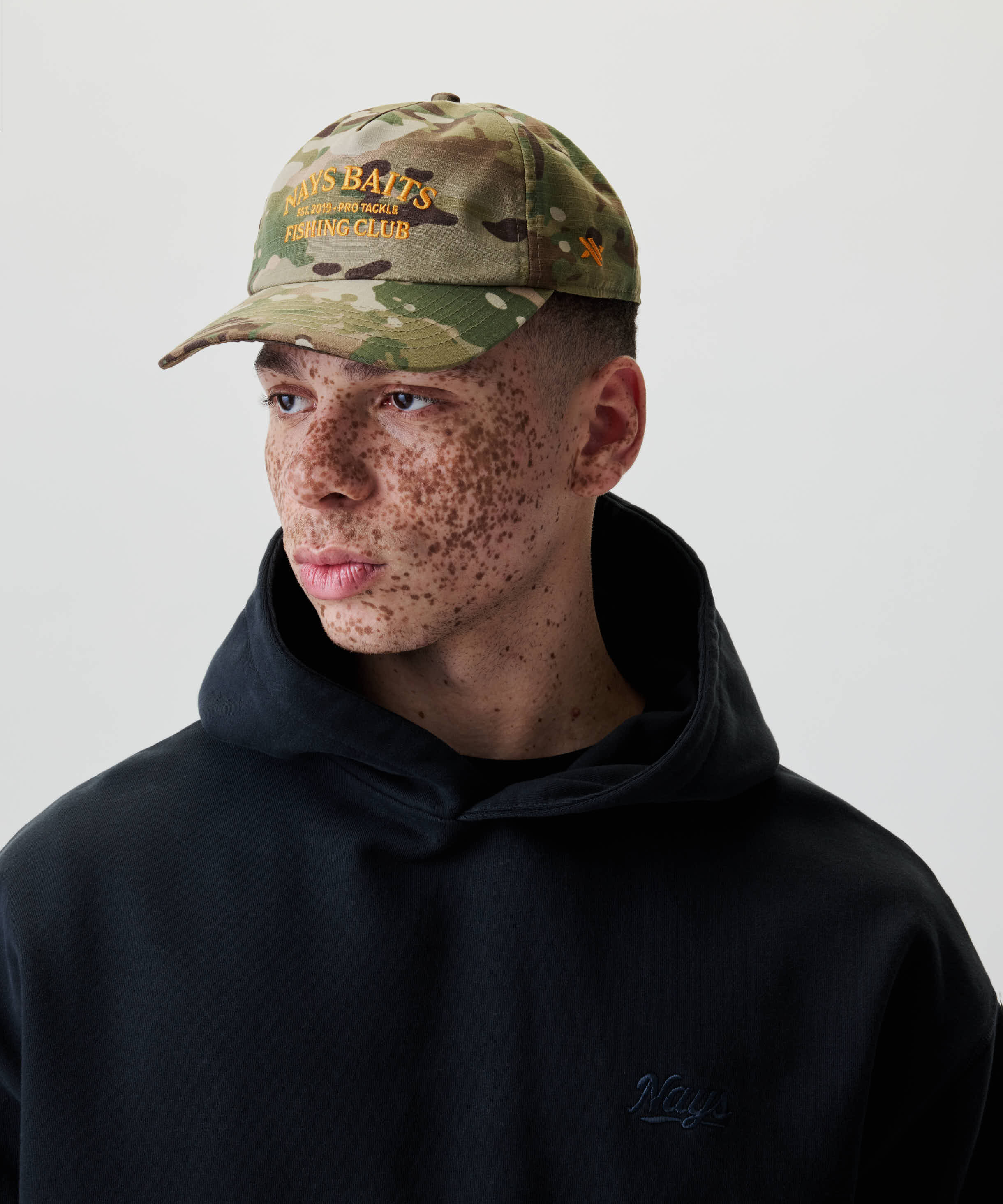 Junger Mann trägt die Nays Fishing Club Cap mit Camo-Muster, orangefarbener Stickerei und Snapback-Verschluss, kombiniert mit schwarzem Hoodie und Nays-Logo.
