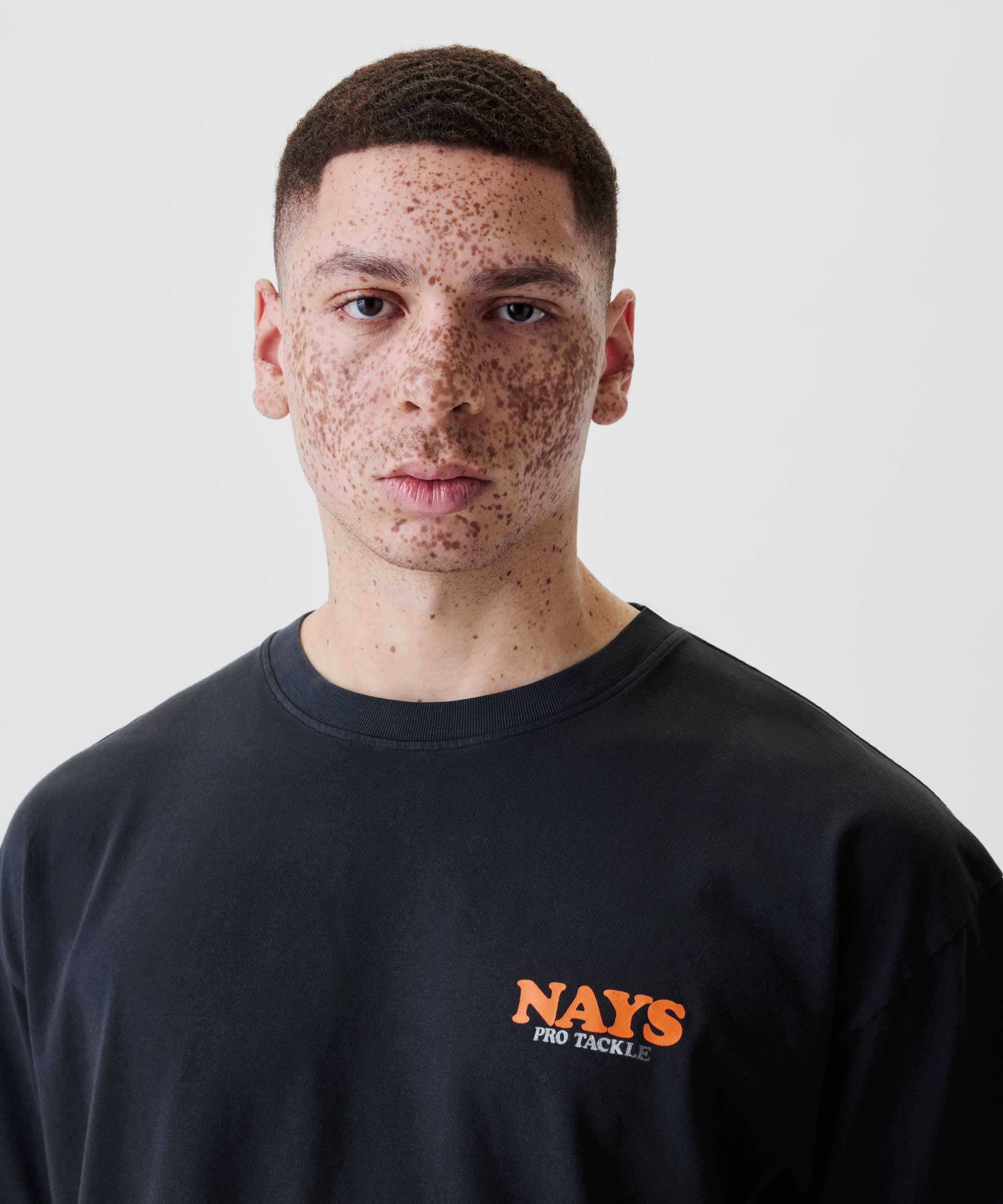 Person mit kurzen dunklen Haaren trägt das Nays College Print Tee in Schwarz mit orange-weißem Brustlogo NAYS PRO TACKLE, vor neutralem Hintergrund.