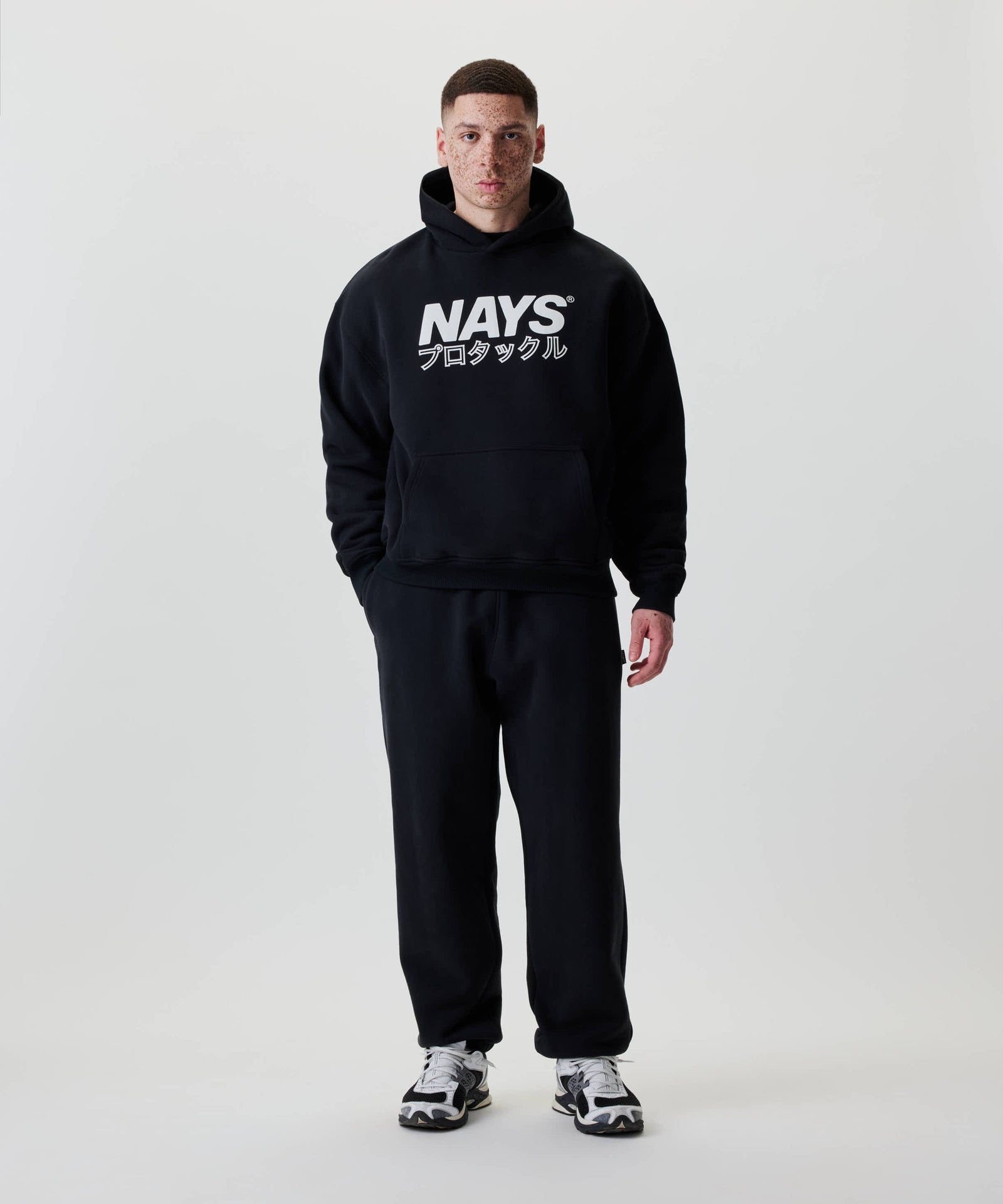 Mann trägt den Nays Understatement Hoodie, schwarz mit weißem Print und japanischen Zeichen, in entspannter Passform, kombiniert mit schwarzen Jogginghosen und sportlichen Sneakern.