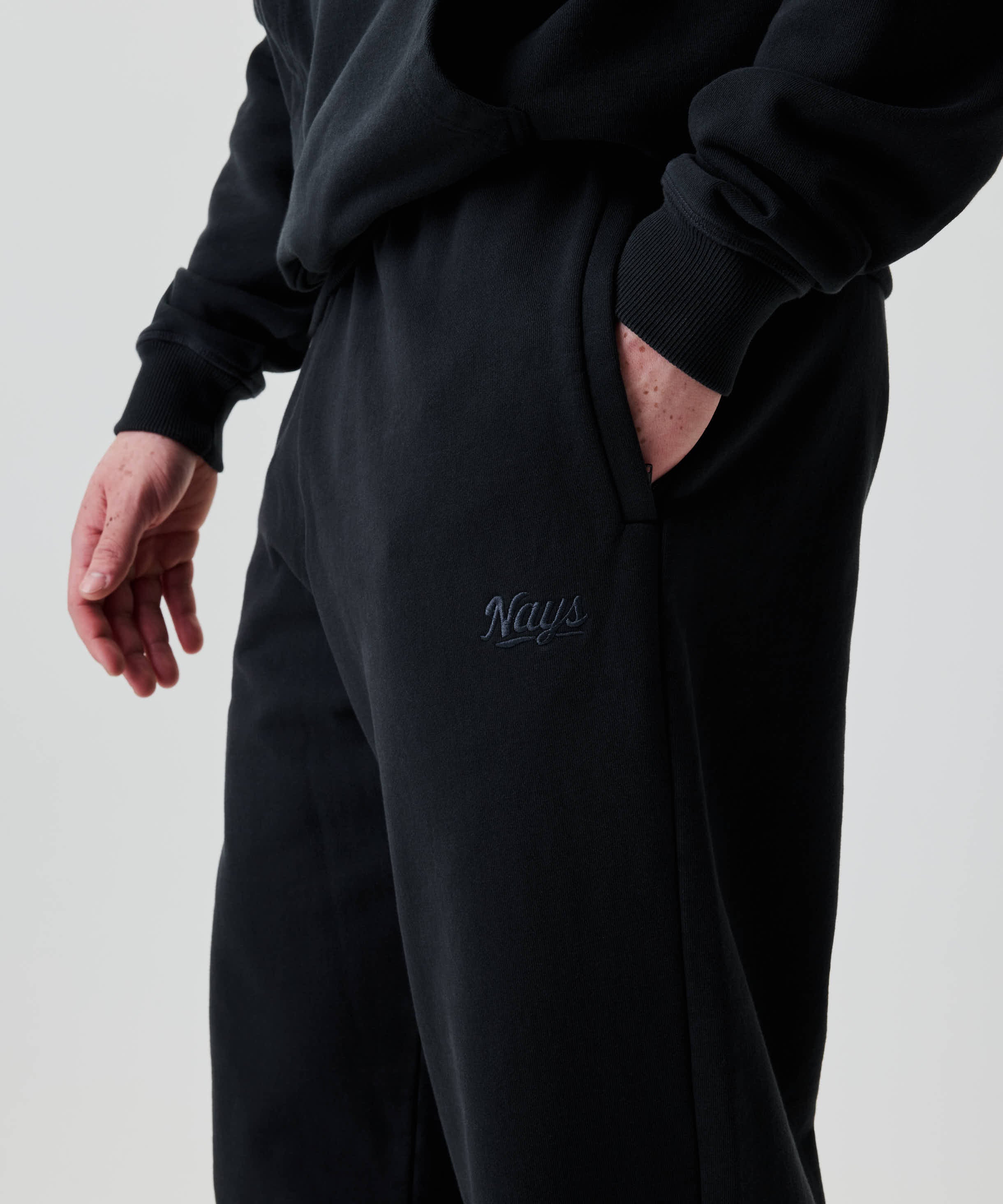 Eine Person trägt Nays Straight Leg Sweatpants mit geradem Bein, seitlichen Taschen und gesticktem Logo Nays auf dem Oberschenkel; eine Hand steckt in der Tasche.