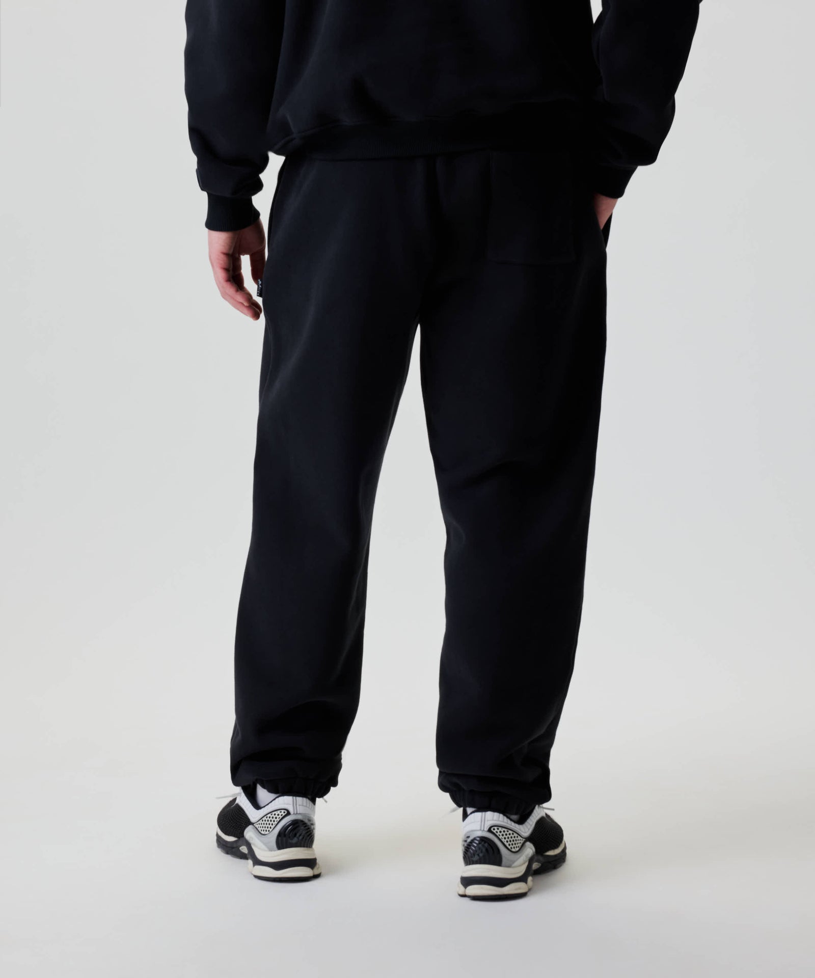 Person trägt Nays Baggy Sweatpants mit elastischen Bündchen und einer rechten Gesäßtasche, kombiniert mit schwarzen Sneakers, linke Hand in der Tasche.