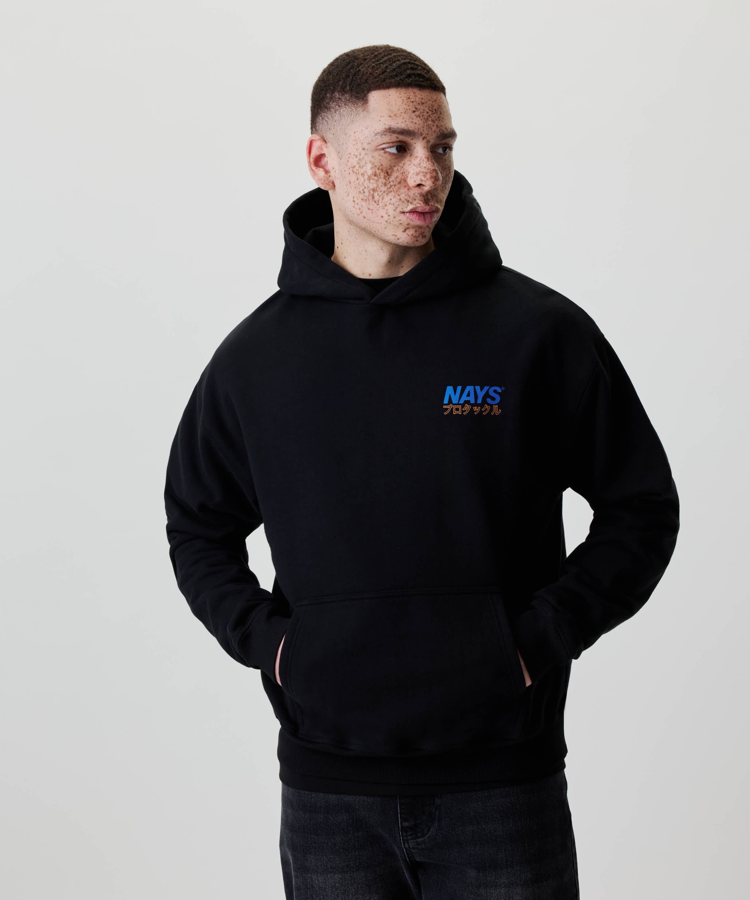 Junger Mann trägt den Nays Pro Tackle International Hoodie mit Brustlogo und Kängurutasche, Hände in der Tasche, blickt zur Seite.