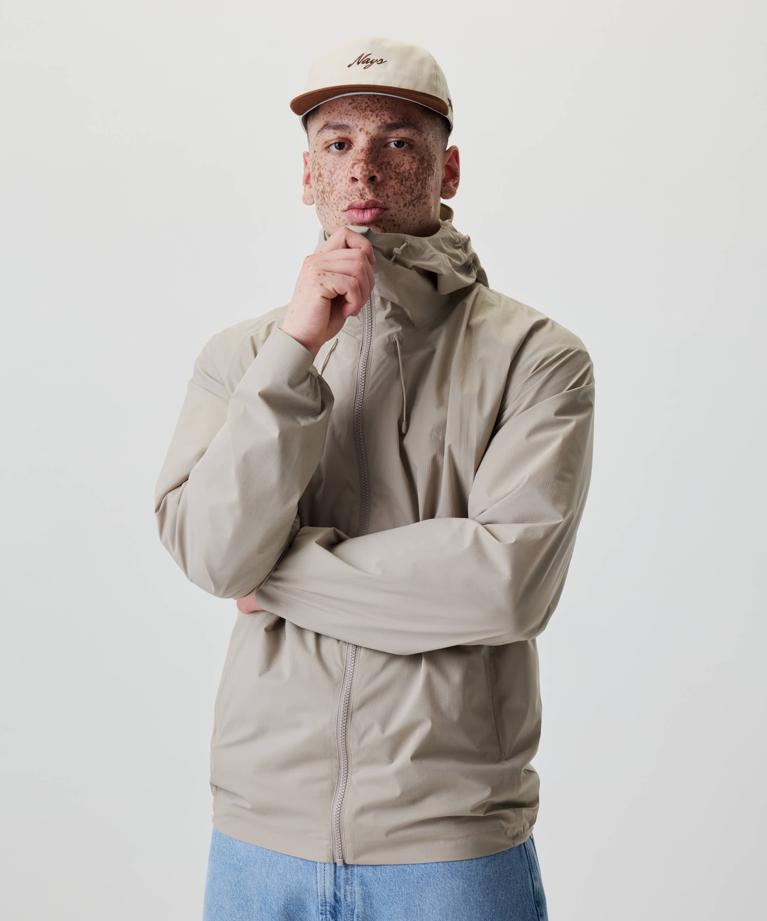 Ein junger Erwachsener trägt die Nays 2L Defend Mono Jacket mit Kapuze und sportlichen Raglanärmeln, kombiniert mit einer passenden Baseballcap.