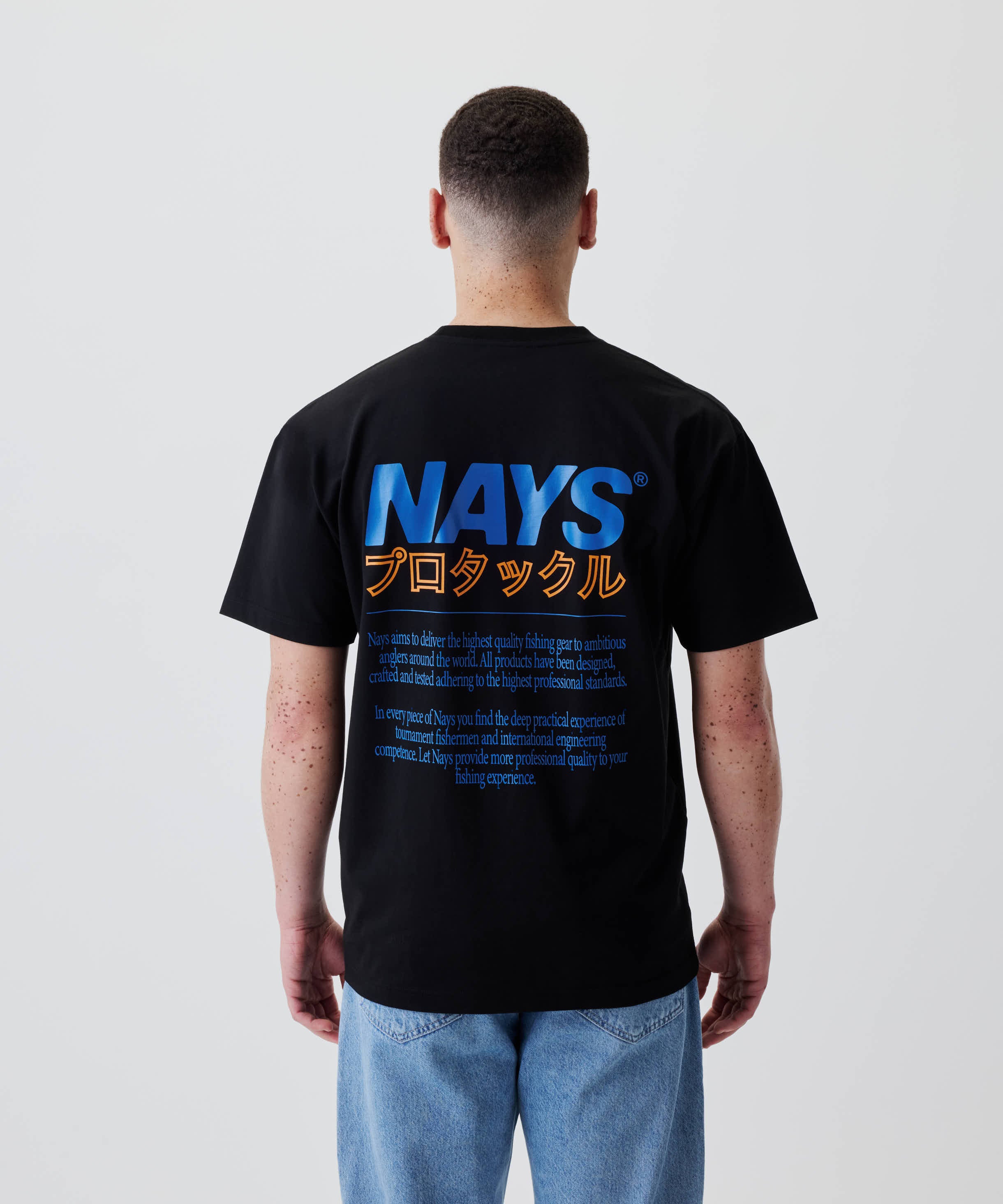 Rückansicht eines Trägers des Nays Pro Tackle International Tee mit großem Rückenprint „NAYS“ und japanischem Text, kombiniert mit blauen Jeans.