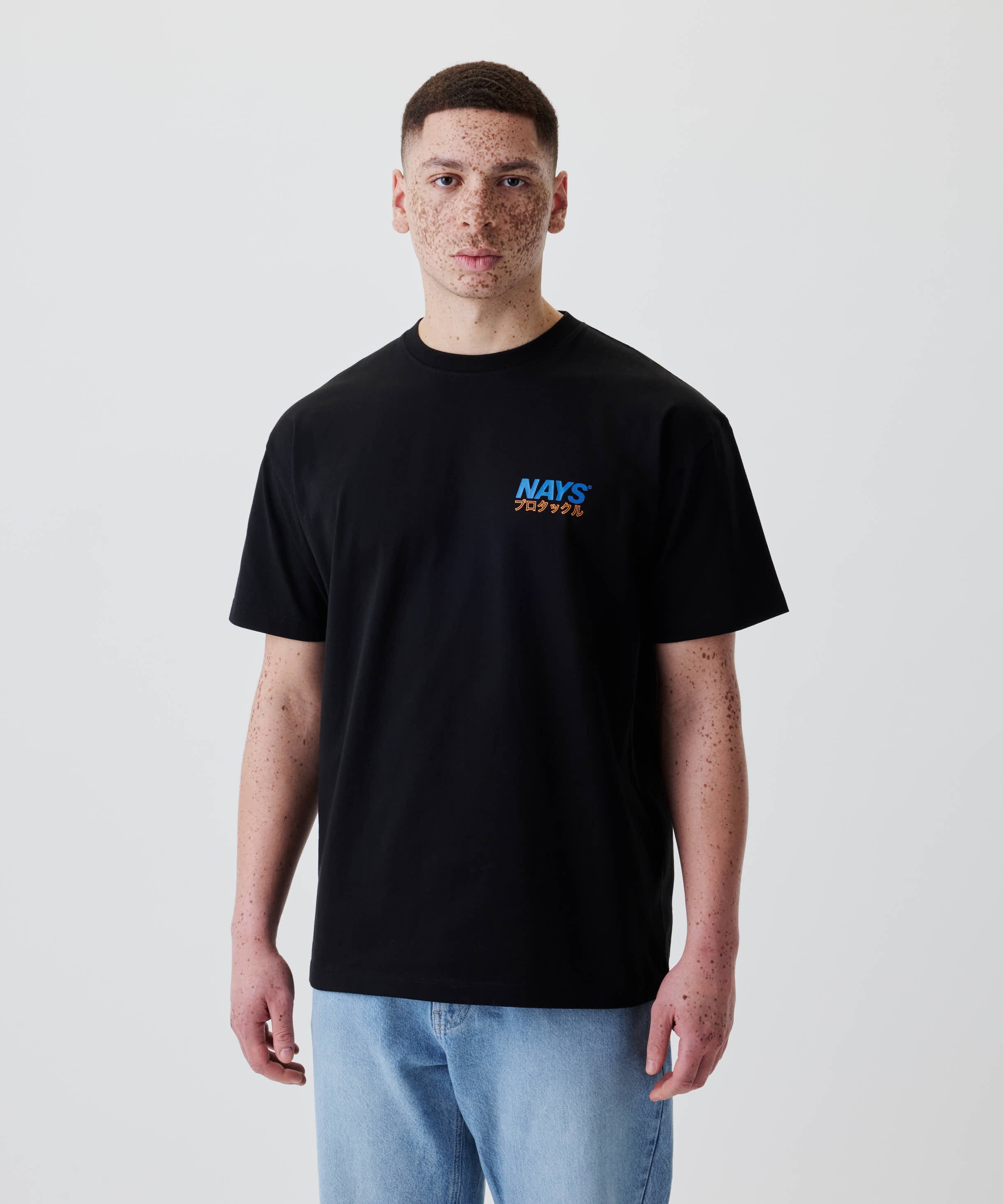 Junger Mann trägt das Nays Pro Tackle International Tee mit kleinem Logo und japanischen Schriftzeichen auf der Brust, kombiniert mit hellblauen Jeans.