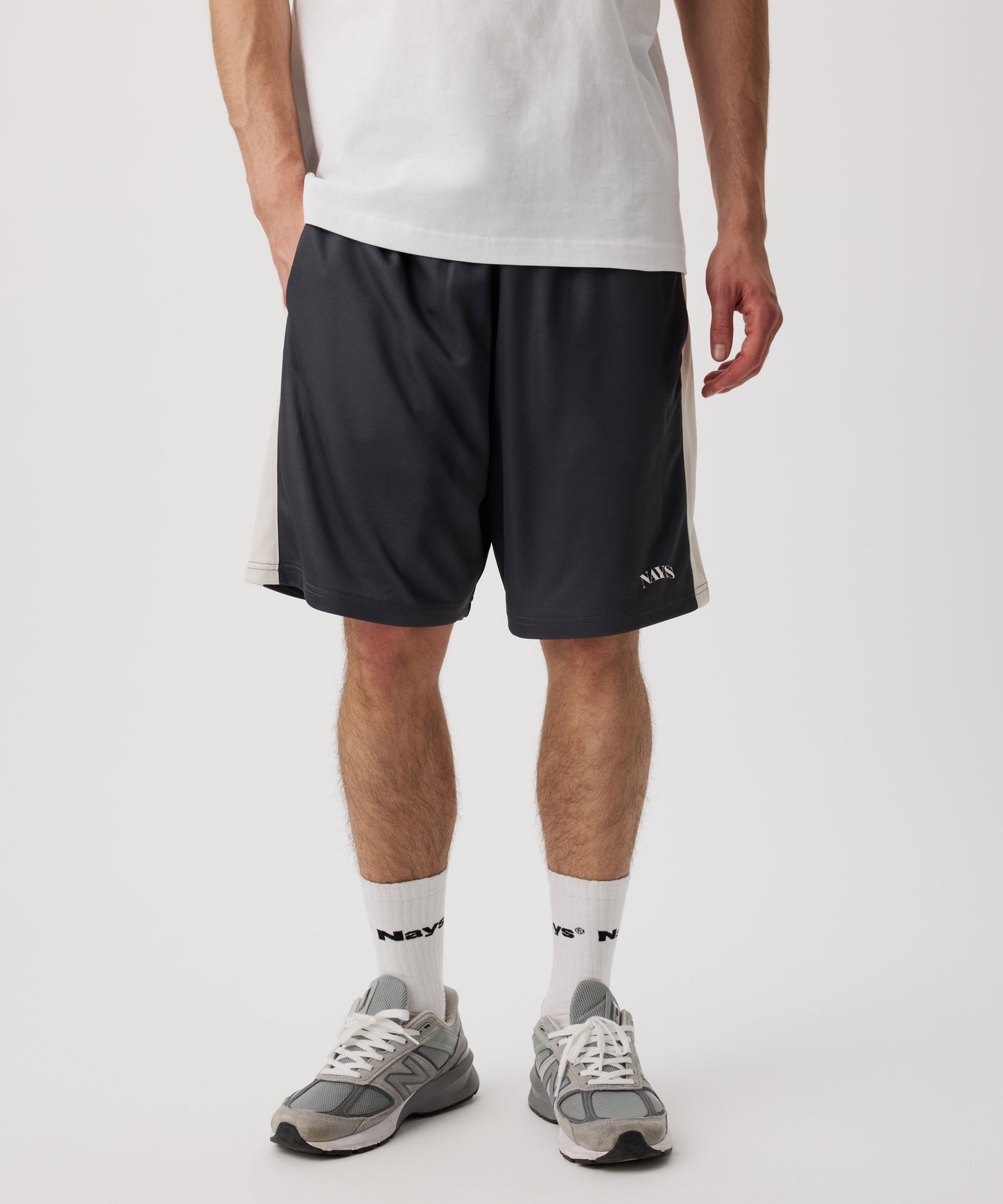 Das Model trägt eine blau-graue-kurze Hose mit weißen Applikationen an den Seiten. Dazu trägt er ein weißes T-Shirt, helle Socken und graue Turnschuhe.