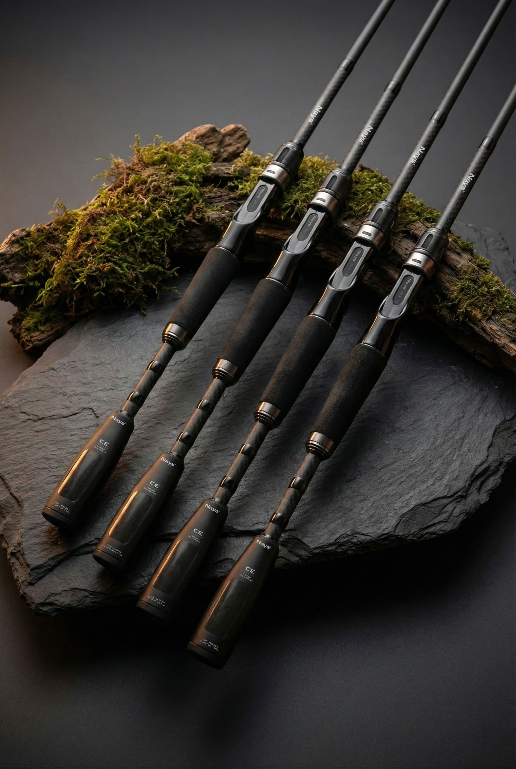 Nays NXT PRO Angelruten im Carbon-Design, schwarze EVA-Griffe, präsentiert auf einer Schieferplatte mit Moos und Holz.