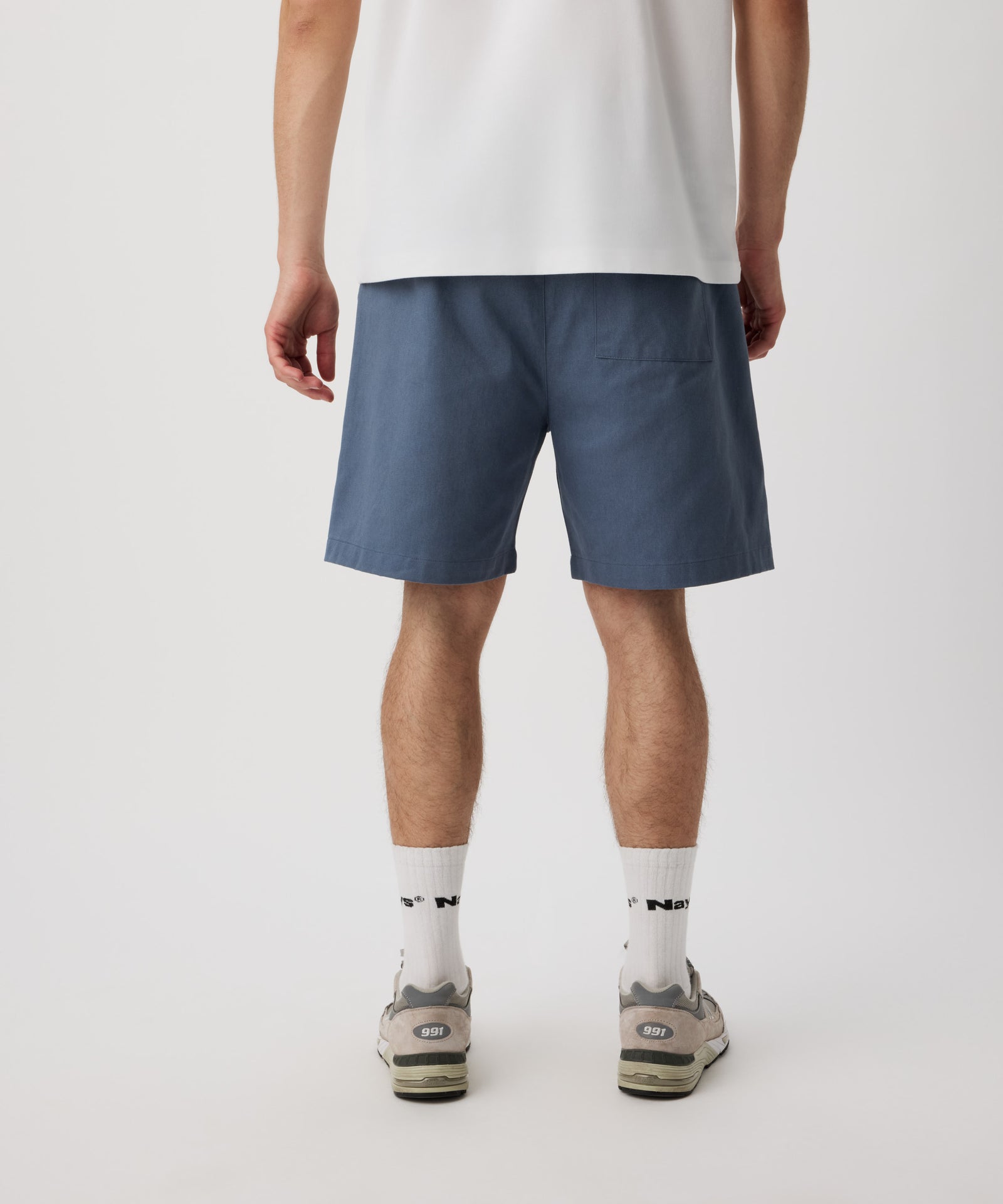 Ein Mann steht mit dem Rücken zur Kamera und trägt ein weißes T-Shirt und eine blaue Shorts. Die kurze Hose verfügt über eine Gesäßtasche. Dazu trägt er weiße Logo Socken und beige-graue Sportschuhe.