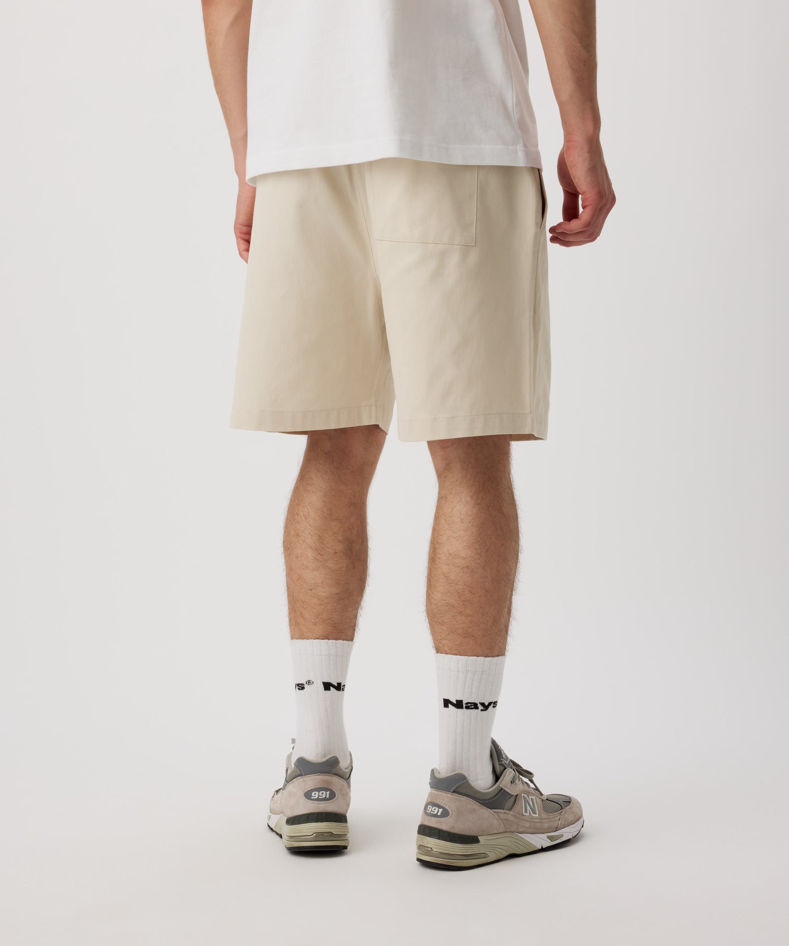 Ein Mann steht mit dem Rücken zur Kamera und trägt ein weißes T-Shirt und eine beige Shorts. Die kurze Hose verfügt über eine Gesäßtasche. Dazu trägt er weiße Logo Socken und Sportschuhe.
