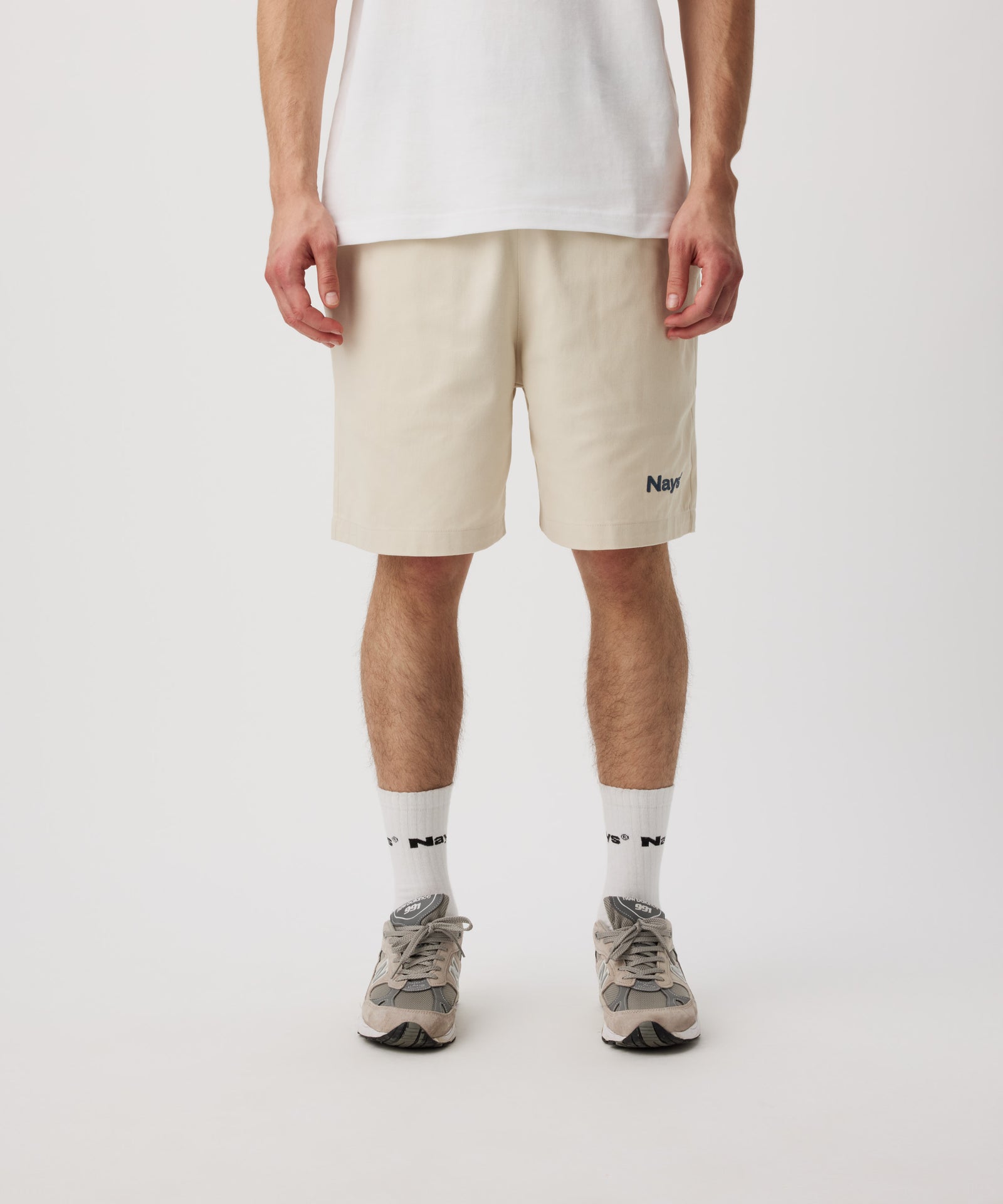 Ein Mann trägt ein weißes T-Shirt, eine beige Shorts mit einem kleinen, grünen Logo auf dem linken Bein. Dazu trägt er weiße Logo Socken und Sportschuhe.