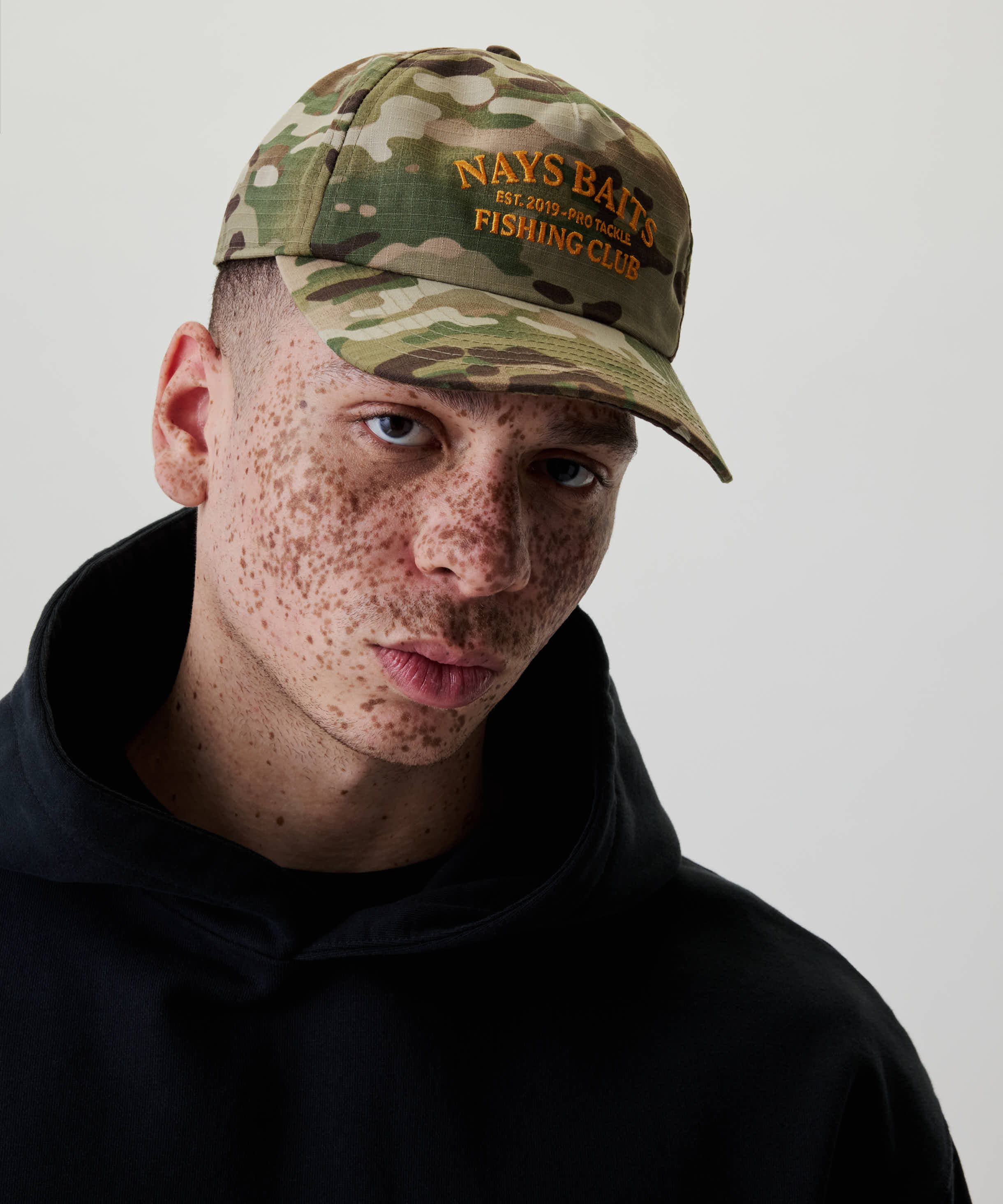Nahaufnahme eines jungen Mannes mit Nays Fishing Club Cap im markanten Camo-Print, bestickt mit orangefarbenem Logo, kombiniert mit schwarzem Hoodie für sportlichen Look.