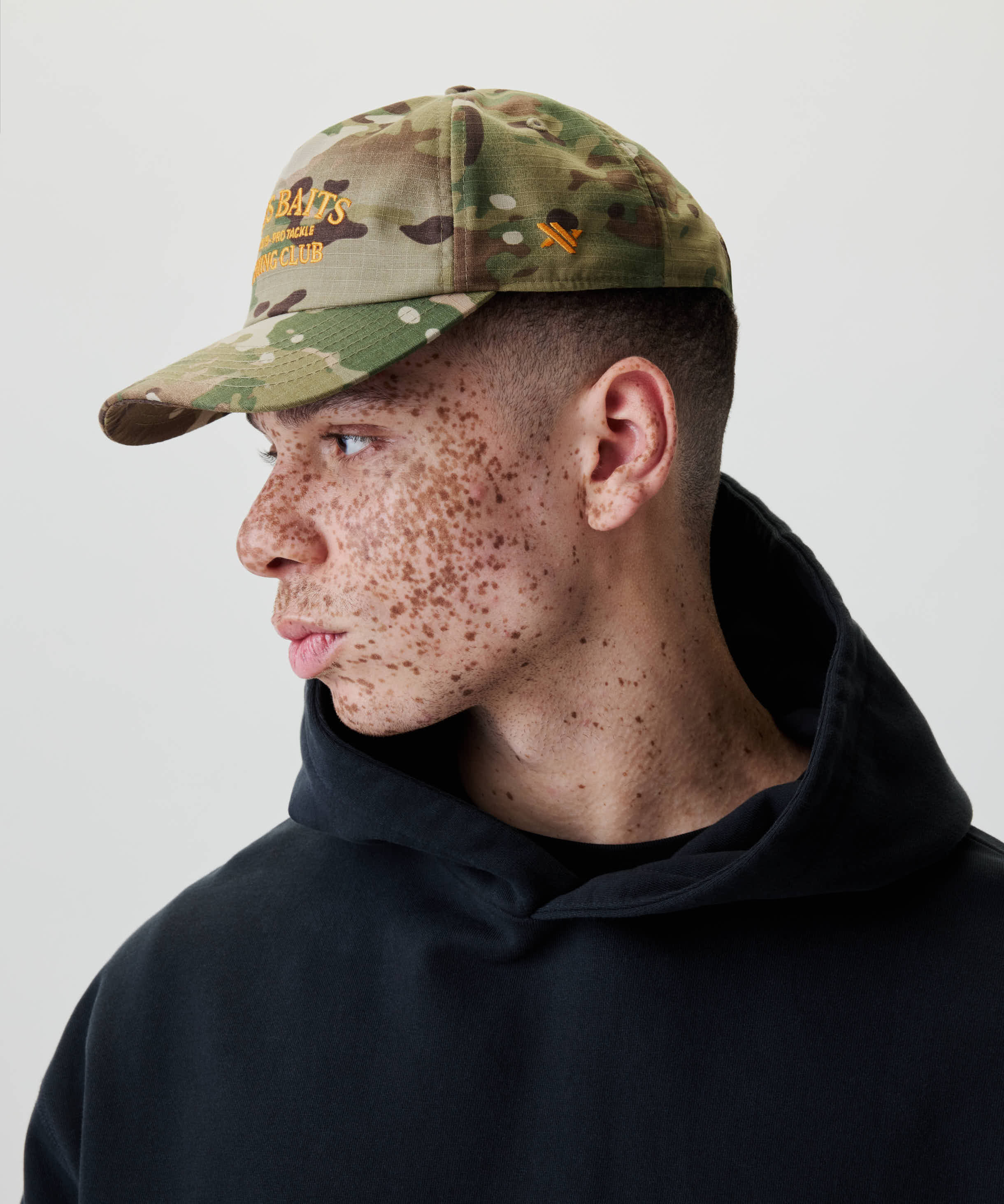 Junge Person im Profil trägt die Nays Fishing Club Cap mit Camouflage-Muster, gelber Stickerei und verstellbarem Snapback für Komfort.