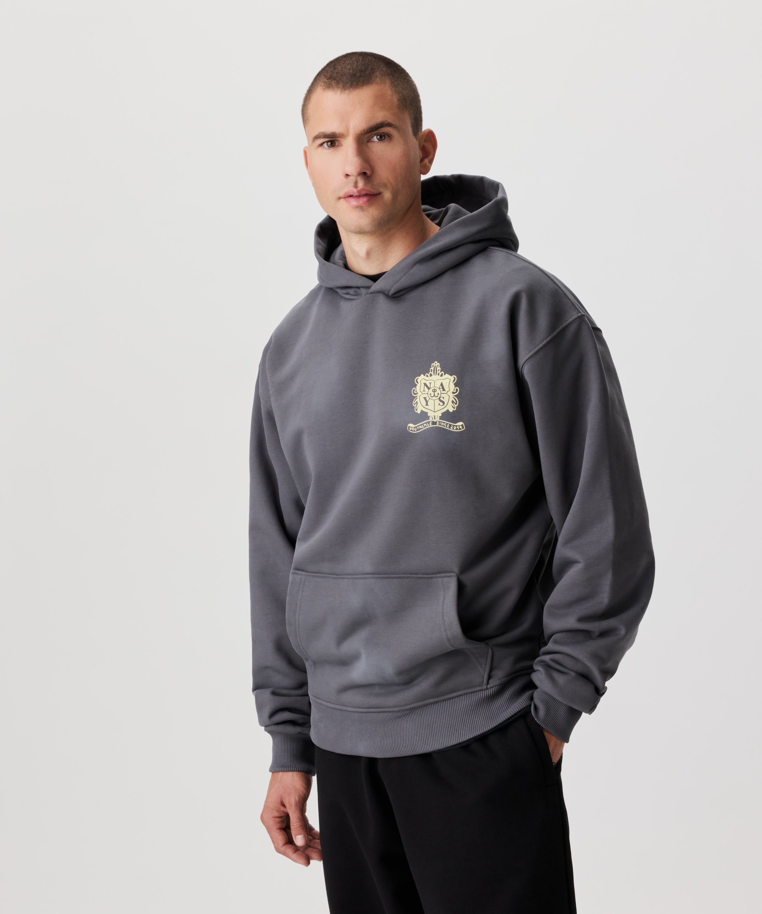 Frontale Ansicht Mann im grauen Kapuzen Sweatshirt und schwarzer Hose. Das Sweatshirt hat einen Druck auf der linken Brust in gelb.