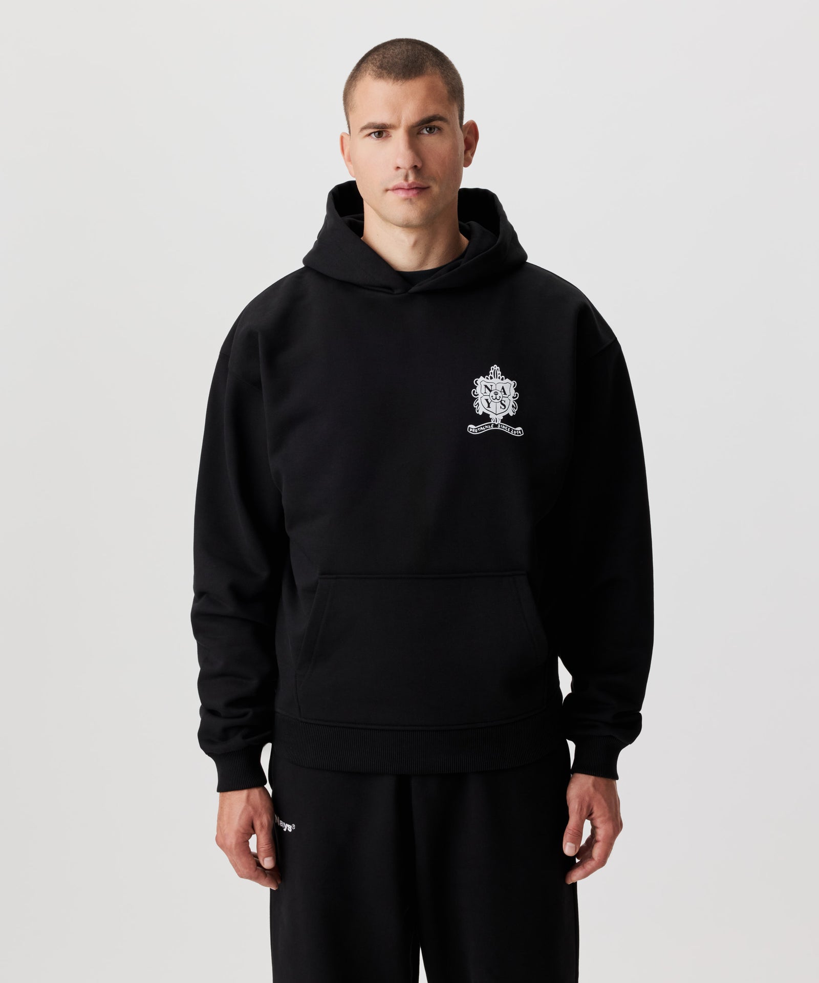 Frontale Ansicht Mann im schwarzen Kapuzen Sweatshirt und schwarzer Hose. Das Sweatshirt hat einen Druck auf der linken Brust in weiß.