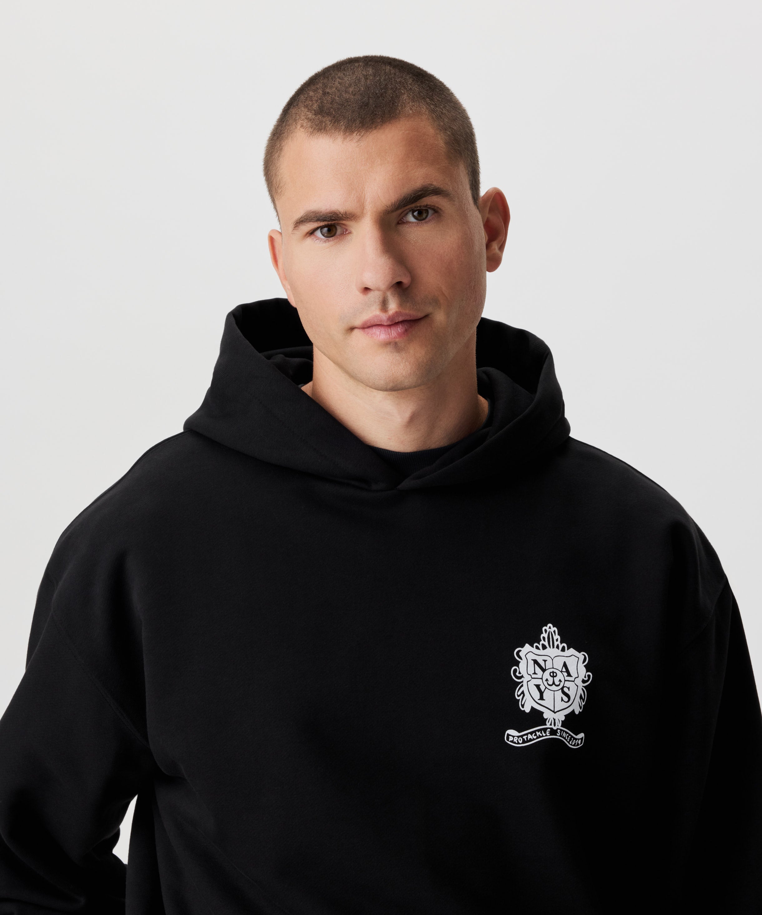 Frontale Ansicht Mann im schwarzen Kapuzen Sweatshirt. Das Sweatshirt hat einen Druck auf der linken Brust in weiß.