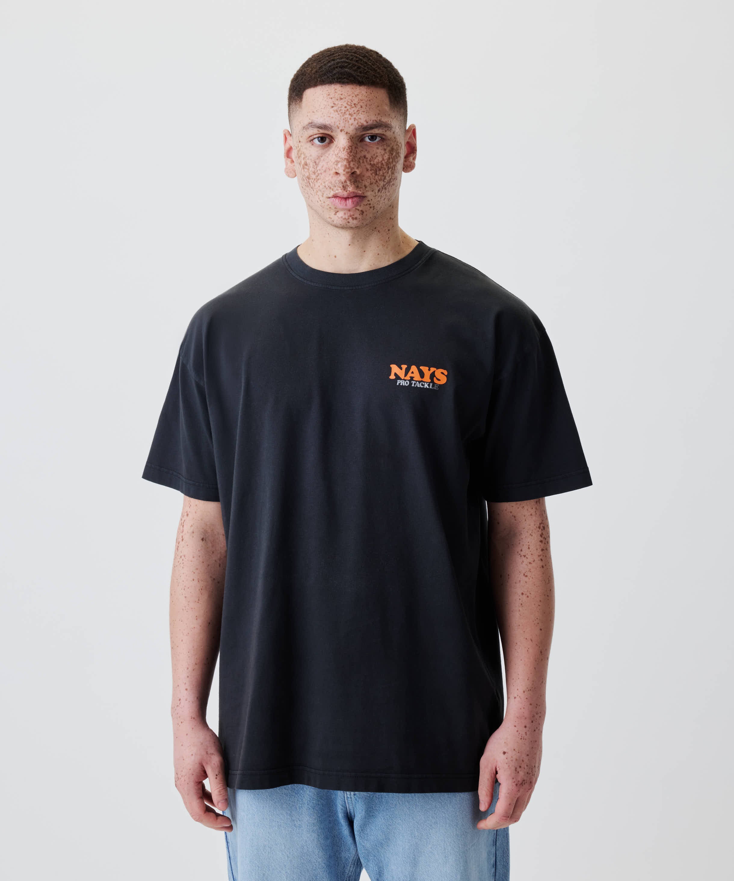 Junger Mann trägt das Nays College Print Tee, ein locker sitzendes schwarzes T-Shirt mit kleinem orangenem Logo NAYS PRO TACKLE auf der Brust.